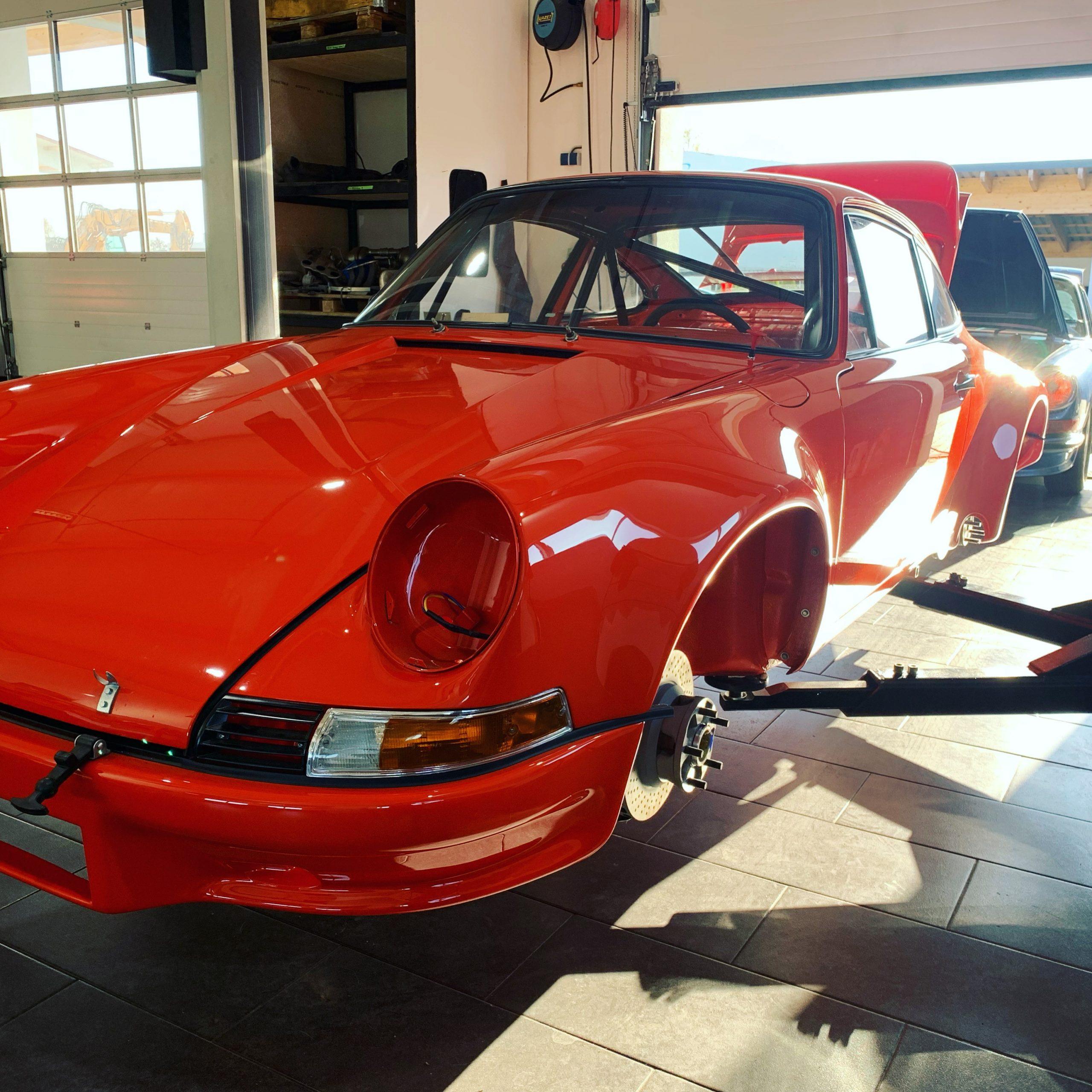 Ein leuchtend roter Oldtimer ohne Räder steht auf einer Hebebühne in einer gut beleuchteten Garage, während das Sonnenlicht hereinfällt und ein anderes Fahrzeug im Hintergrund teilweise sichtbar ist.
