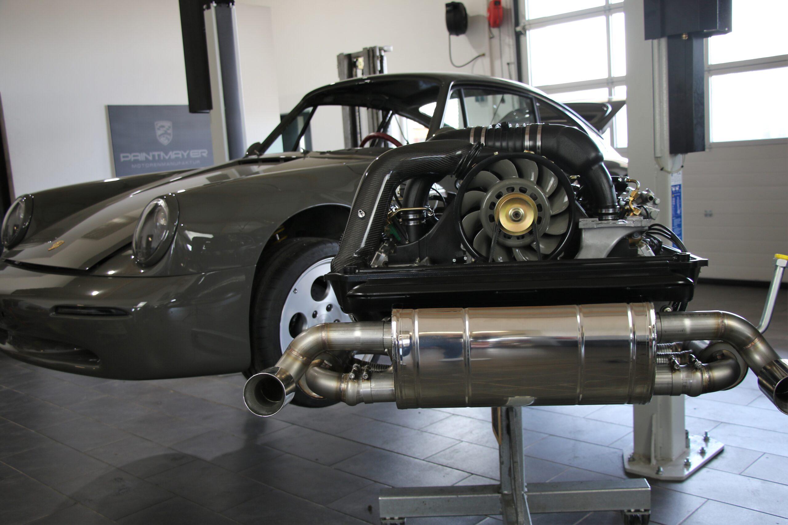 Ein klassischer Porsche 911 mit ausgebautem Motor liegt auf einer Hebebühne in einer Kfz-Werkstatt; im Vordergrund sind der ausgebaute Motor und die Auspuffanlage zu sehen.