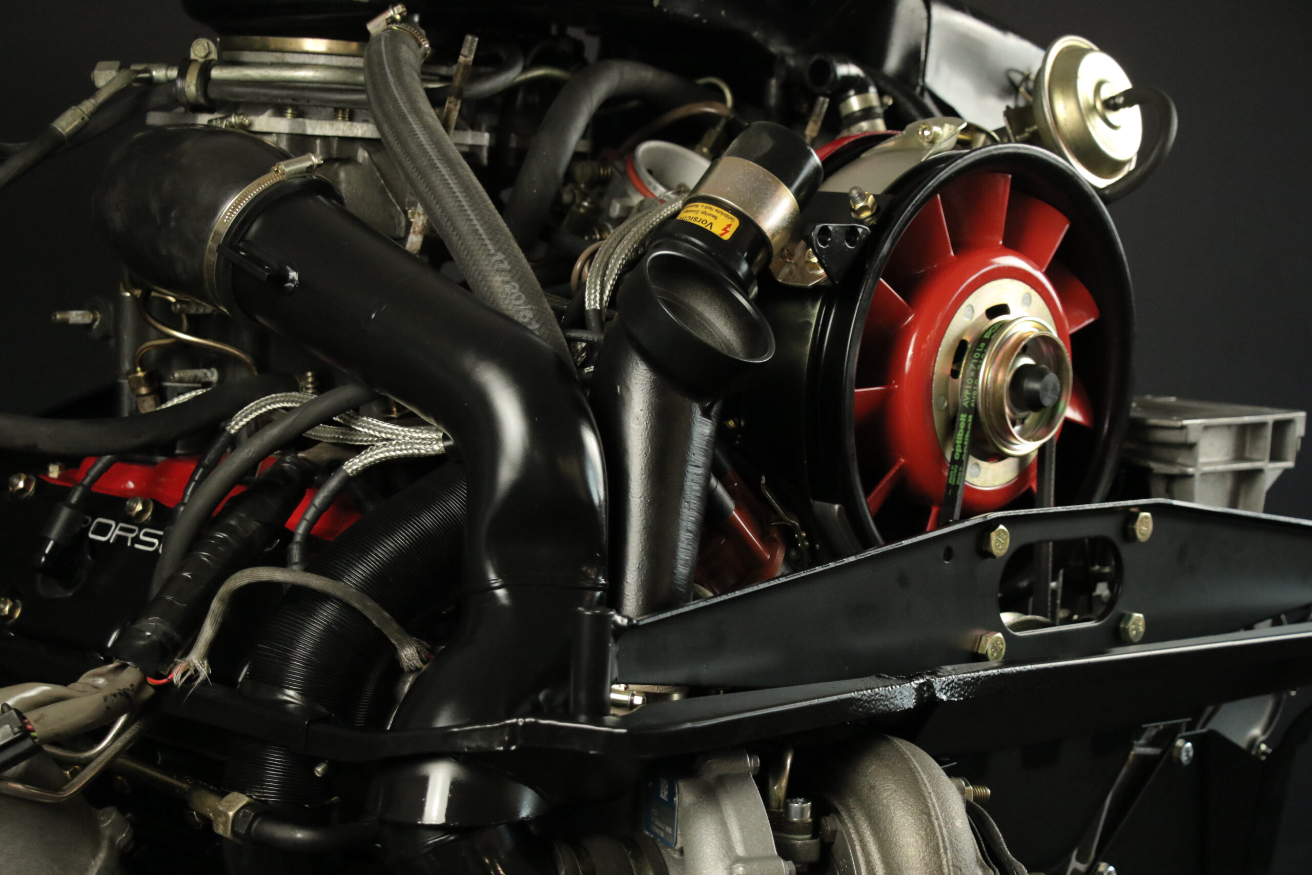 Nahaufnahme eines Oldtimer-Motors mit sichtbaren schwarzen Schläuchen, Metallrohren und einem auffälligen roten Kühlerlüftergehäuse. Die Teile sind sauber und gut gepflegt und stehen vor einem dunklen Hintergrund.