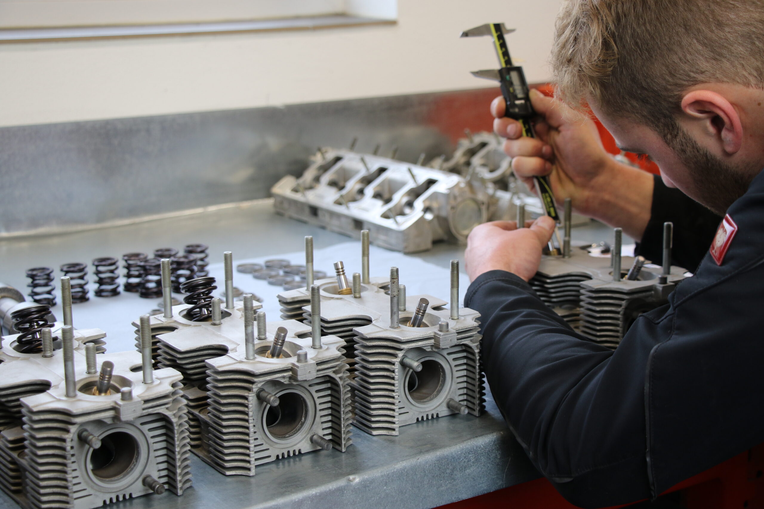Eine Person misst mit einem Messschieber ein Motorteil aus Metall auf einer Werkbank, die mit ähnlichen Teilen und kleinen Metallfedern ausgelegt ist. Im Hintergrund sind weitere Motorenteile angeordnet.