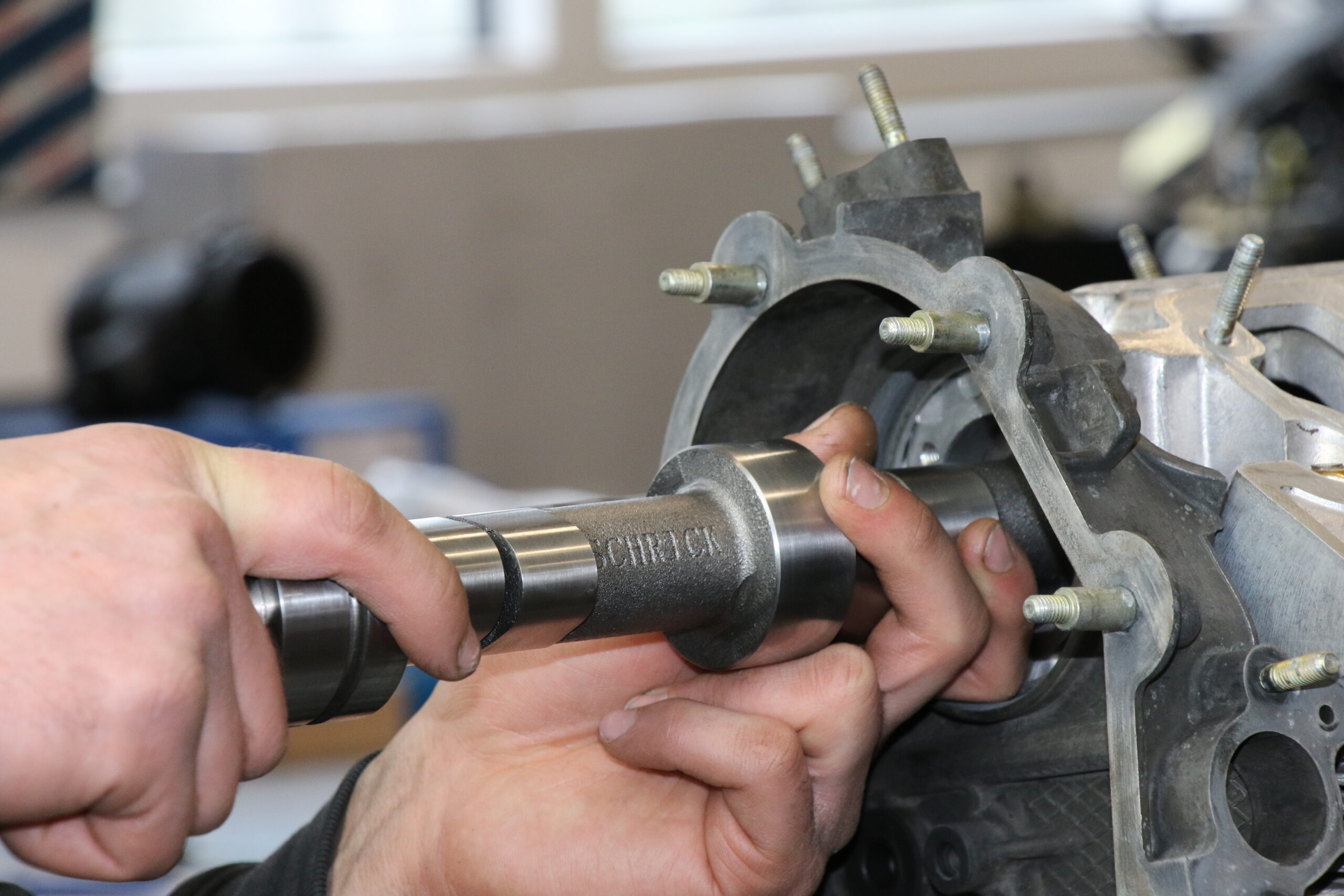 Eine Person setzt eine Nockenwelle aus Metall in einen Motorblock eines Autos ein, wobei sie beide Hände benutzt, um das Teil in die richtige Position zu bringen. Der Motorblock und die Schrauben sind sichtbar, der Hintergrund ist unscharf.