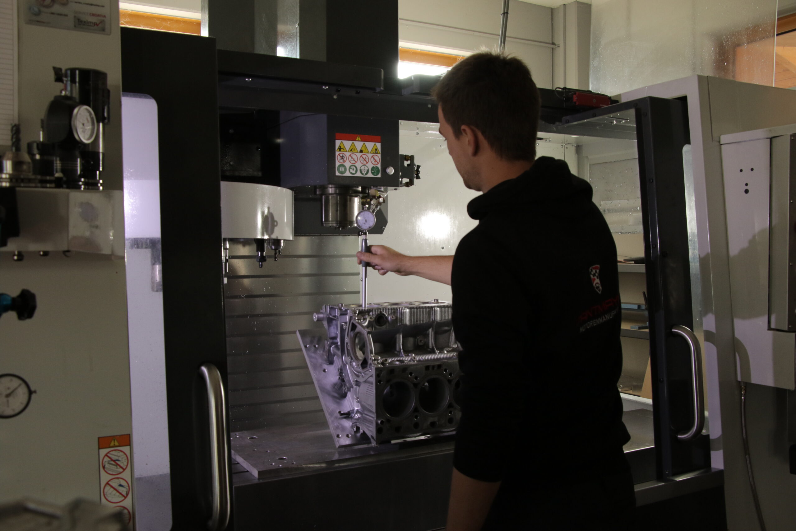 Eine Person bedient eine CNC-Maschine und stellt ein Werkzeug in der Maschine ein, während sie in einer Werkstatt an einem großen Metallmotorblock arbeitet.