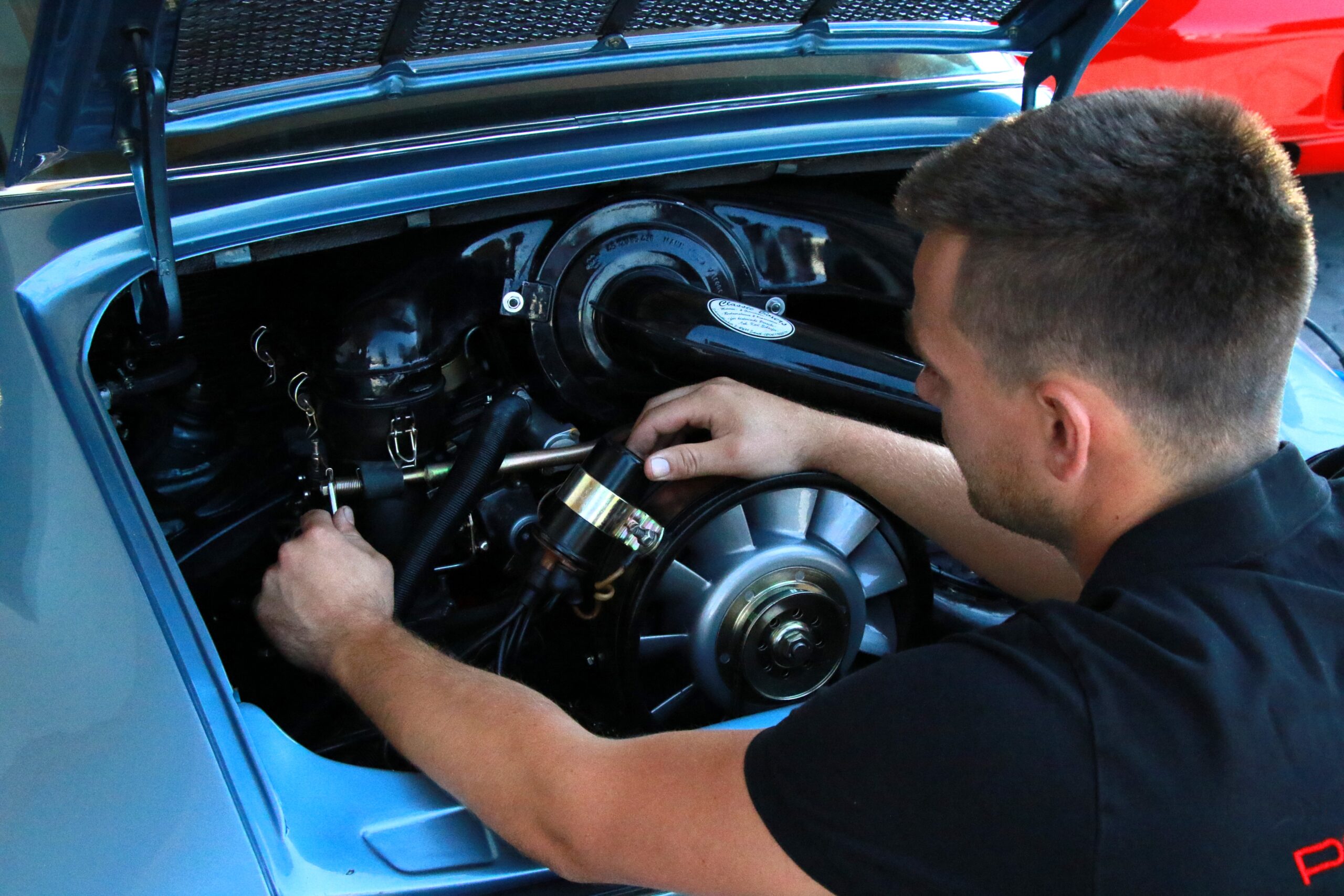 Ein Mann arbeitet bei geöffneter Motorhaube am Motor eines Oldtimers und benutzt seine Hände, um Motorteile einzustellen oder zu reparieren. Der Motor ist sauber und gut gewartet.