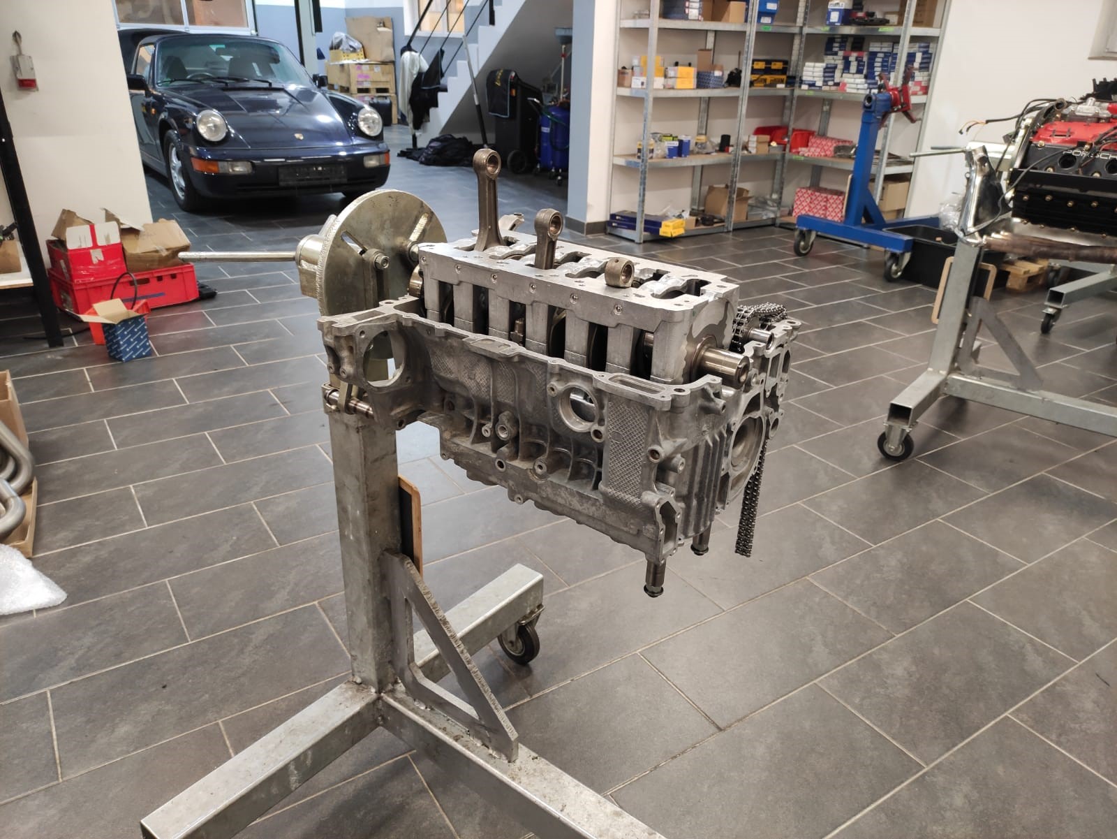 Ein zerlegter Motorblock, montiert auf einem Ständer in einer Werkstatt mit Werkzeugen, Regalen und einem Oldtimer im Hintergrund.