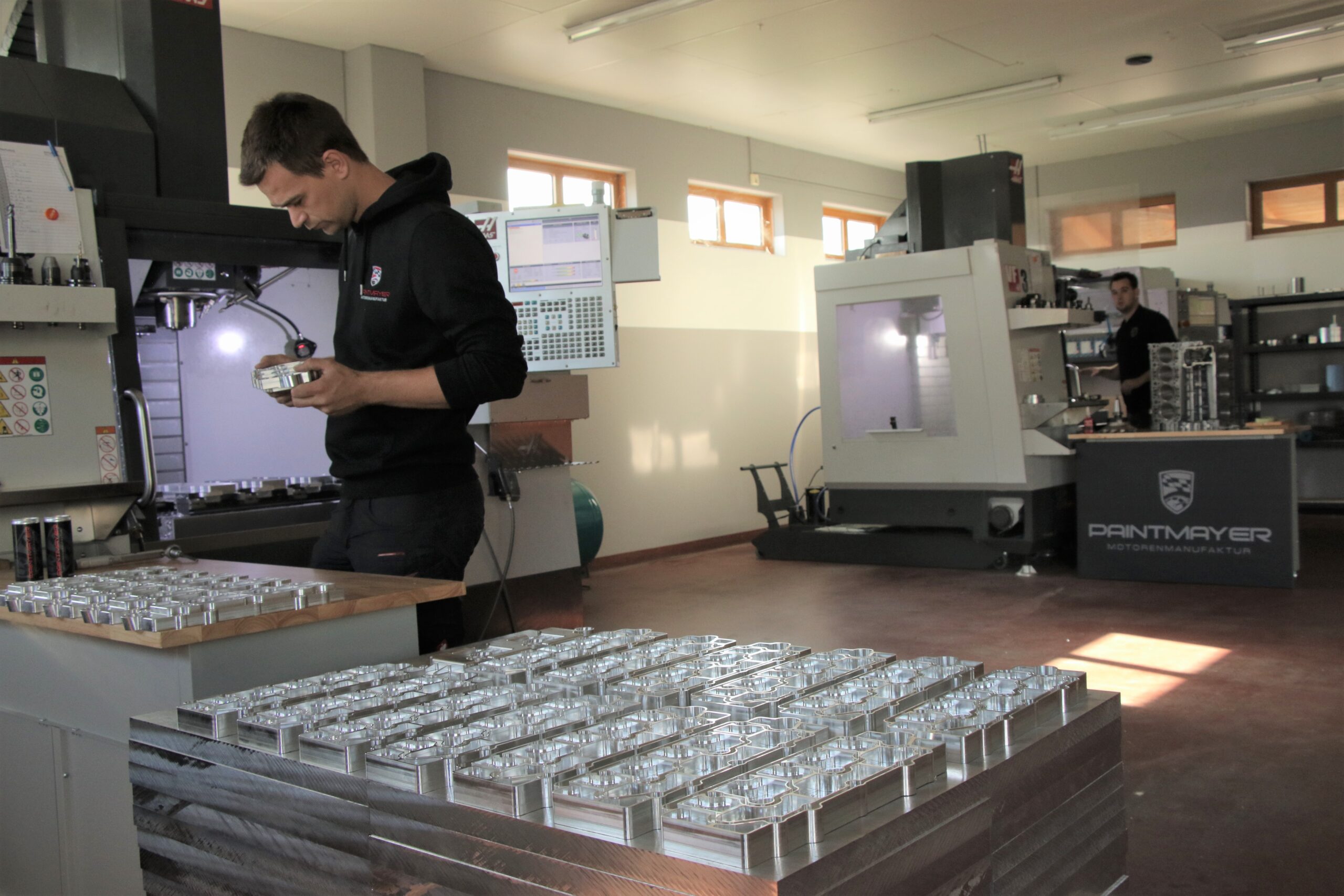 Ein Mann prüft ein Metallteil in einer Werkstatt, die mit ordentlich angeordneten Metallteilen gefüllt ist. Eine andere Person arbeitet an einer Maschine im Hintergrund. Der Arbeitsplatz ist gut beleuchtet, mit modernen Geräten und Computerbildschirmen ausgestattet.