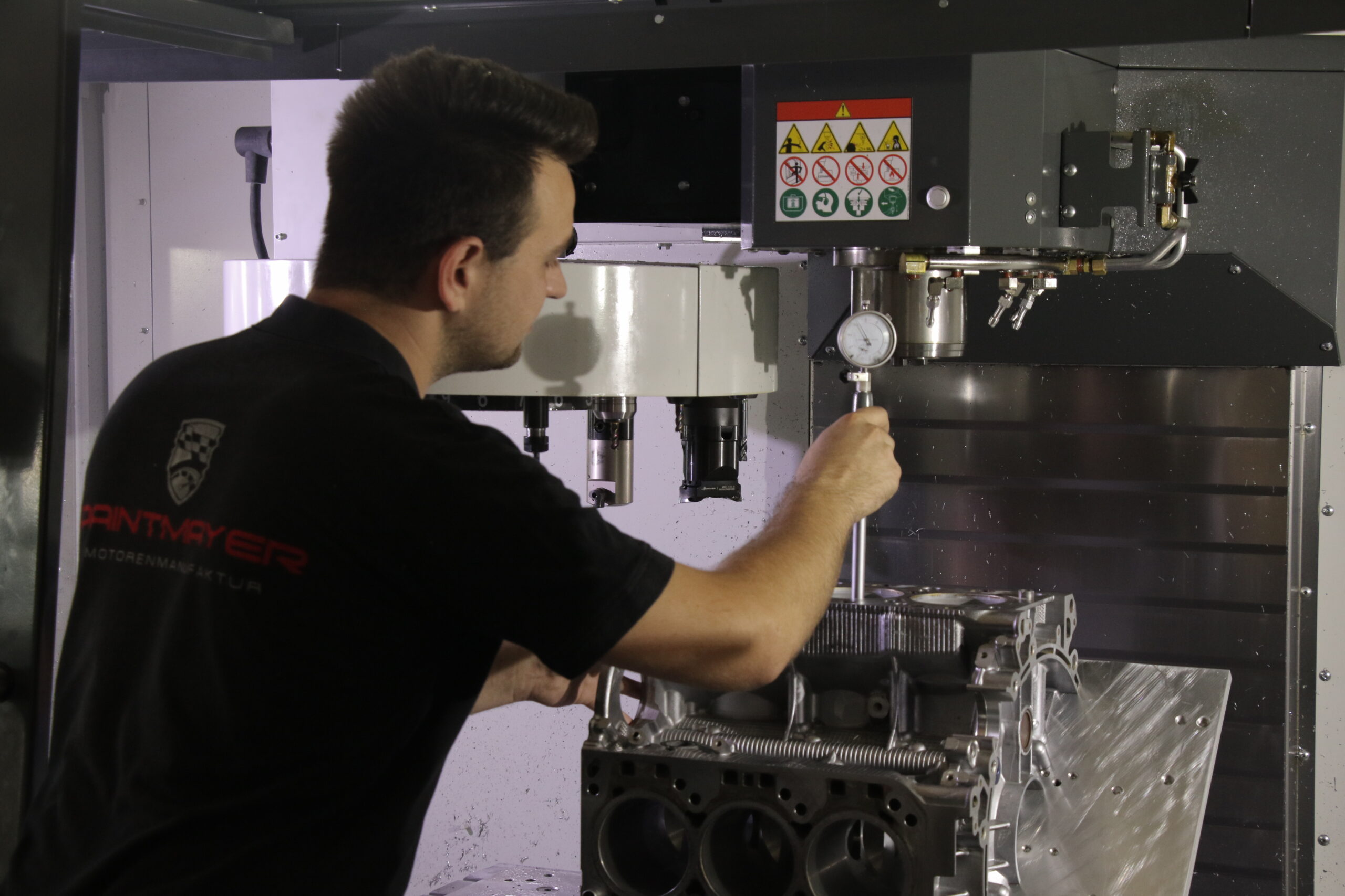 Ein Mann bedient Maschinen und misst mit einer Messuhr einen Metallmotorblock in einer CNC-Maschine. Er trägt ein schwarzes Hemd mit einem Logo auf dem Ärmel und konzentriert sich auf seine präzise Arbeit.