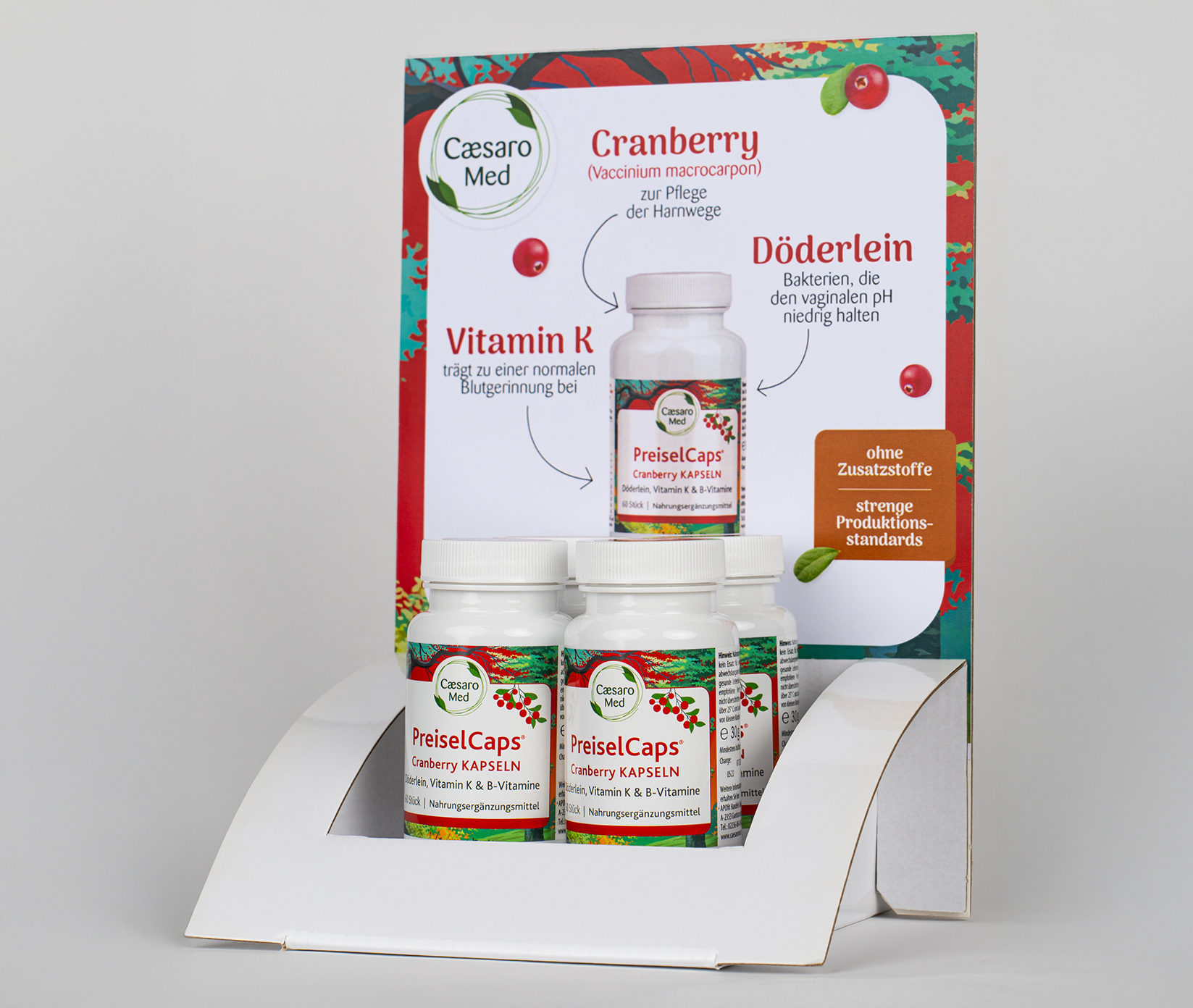 Apofit_Steher_2©invisions Kopie Drei weiße Flaschen Ceasaro Med PreiselCaps Cranberry-Kapseln sind in einem weißen Ständer ausgestellt. Dahinter befindet sich ein Informationsdisplay mit Bildern des Produkts und Texten über Cranberry, Vitamin K und Döderlein.