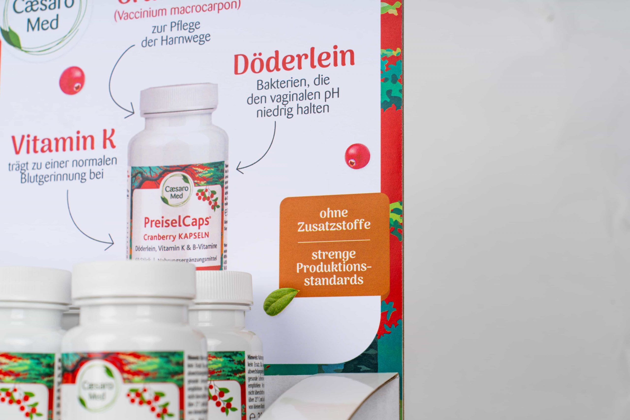 Nahaufnahme eines Produktdisplays für Caesar Med PreiselCaps Cranberry Kapseln mit Ergänzungsflaschen und deutschem Text, der Merkmale wie "ohne Zusatzstoffe" und "strenge Produktionsstandards" hervorhebt.