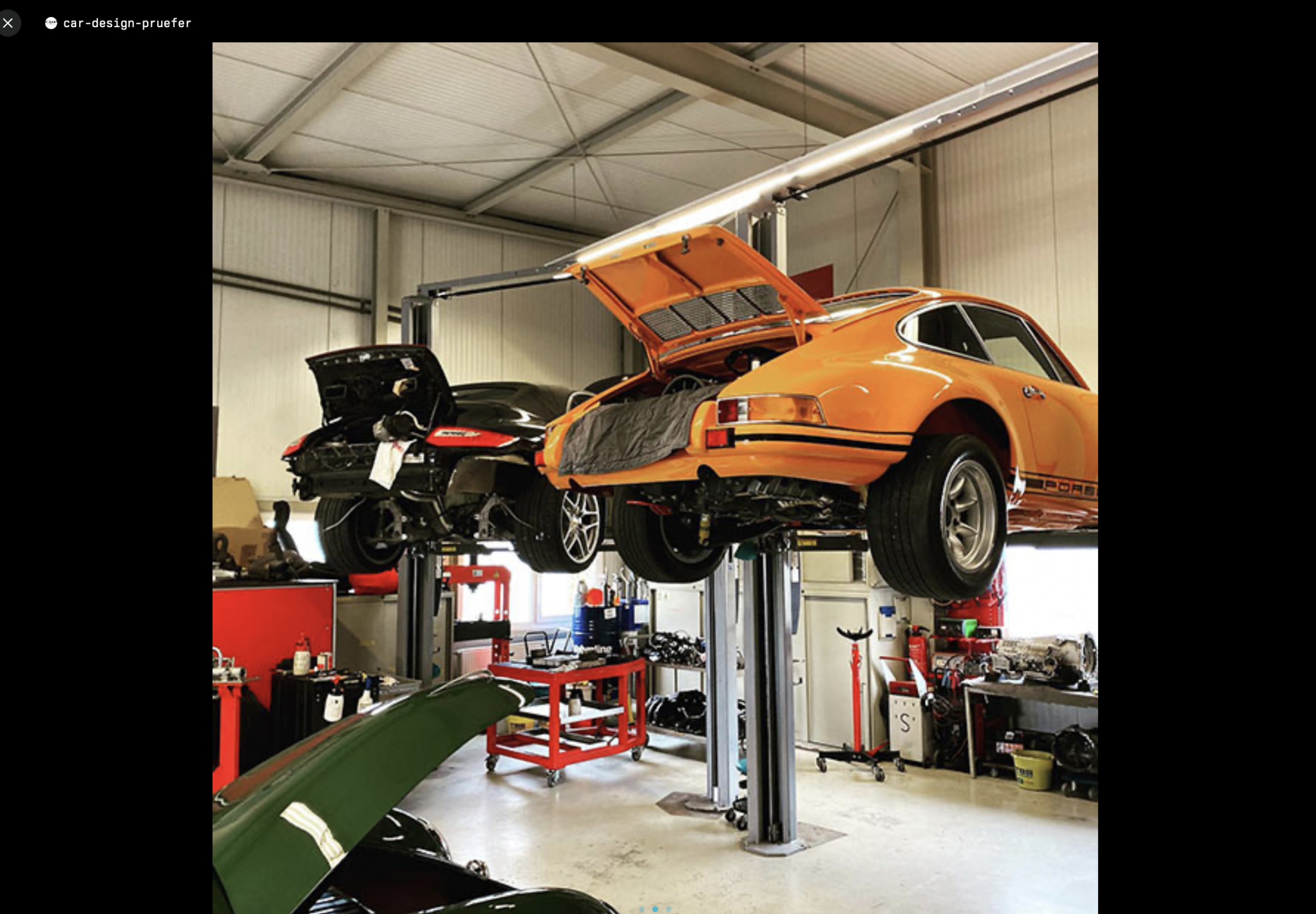 Zwei Oldtimer, darunter ein orangefarbener Porsche mit offenem Kofferraum, stehen auf Hebebühnen in einer gut beleuchteten Autowerkstatt voller Werkzeuge und Geräte.