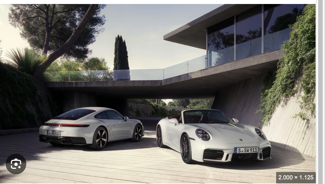 Zwei weiße Porsche 911 Sportwagen, ein Coupé und ein Cabrio, stehen auf einer modernen Auffahrt vor einem modernen Haus mit Glasgeländer und umgeben von Grünflächen.