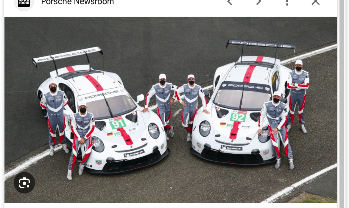 Zwei weiße Porsche-Rennwagen mit roten und grauen Akzenten sind nebeneinander auf einer Rennstrecke geparkt. Acht Teammitglieder in passenden Rennuniformen und Masken stehen neben den Autos und posieren für ein Gruppenfoto.