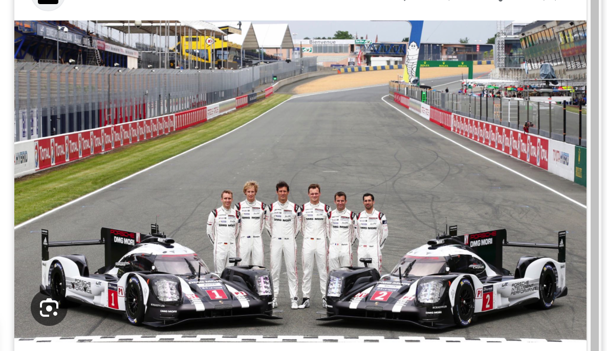 Sechs Rennfahrer in weißen Rennanzügen stehen auf einer Rennstrecke zwischen zwei schwarz-weißen Porsche-Rennwagen, im Hintergrund sind Tribünen und Renninfrastruktur zu sehen.