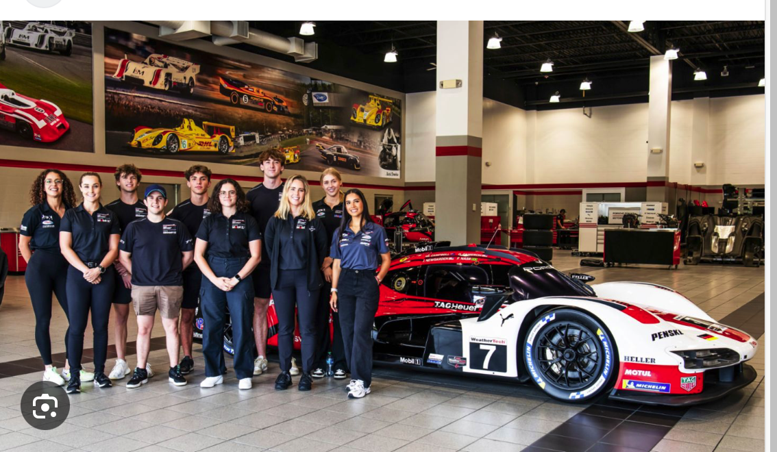 Eine Gruppe von neun Personen in passenden Team-Shirts posiert neben einem schwarz-weiß-roten Rennwagen mit der Aufschrift 7 in einem Ausstellungsraum mit Rennplakaten und Autos im Hintergrund.