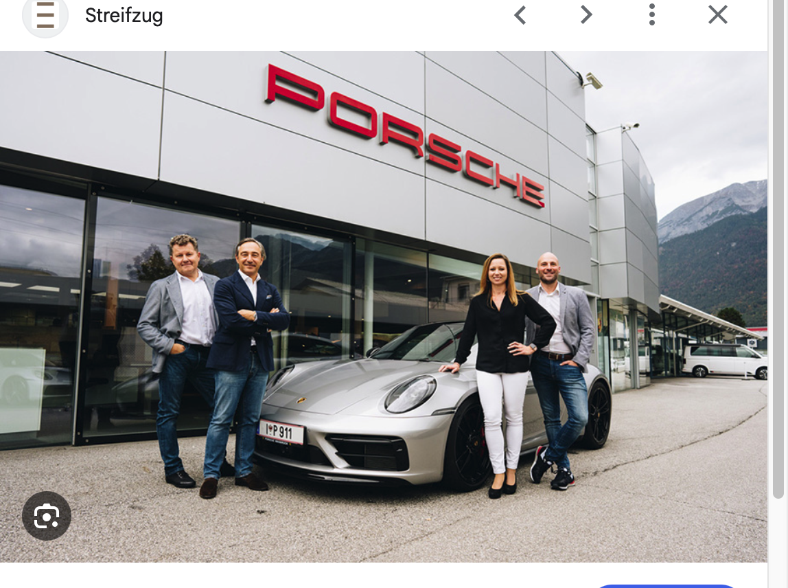 Vier Personen stehen vor einem silbernen Porsche vor dem Gebäude eines modernen Porsche-Händlers, im Hintergrund sind die Berge zu sehen.