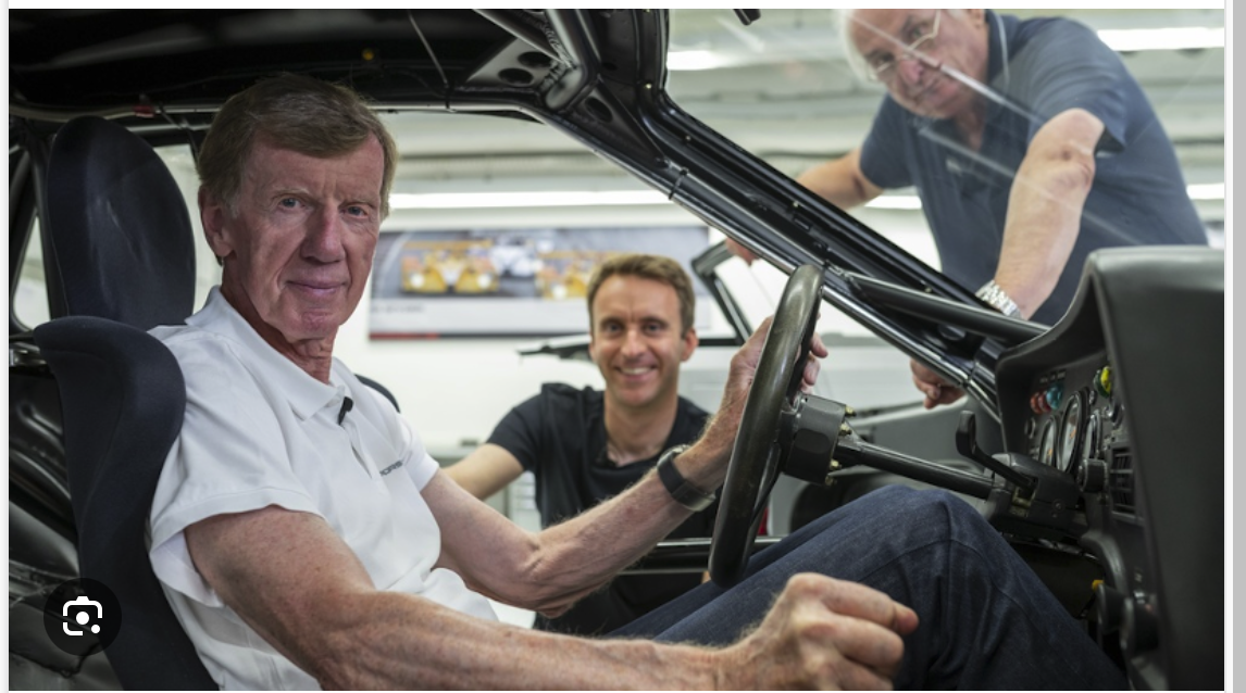 Ein älterer Mann sitzt auf dem Fahrersitz eines Autos und lächelt in die Kamera. Vor dem Auto stehen zwei Männer, die ebenfalls lächeln, in einer gut beleuchteten Garage oder Werkstatt.