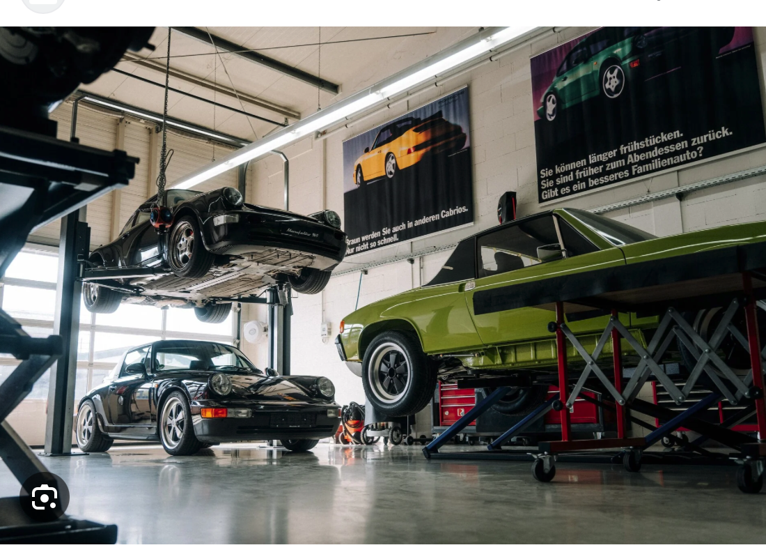 Eine Autowerkstatt mit zwei Porsche-Klassikern auf einer Hebebühne und einem weiteren schwarzen Porsche, der auf dem Boden geparkt ist. In der gut beleuchteten Werkstatt hängen Plakate von Oldtimern an der Wand.
