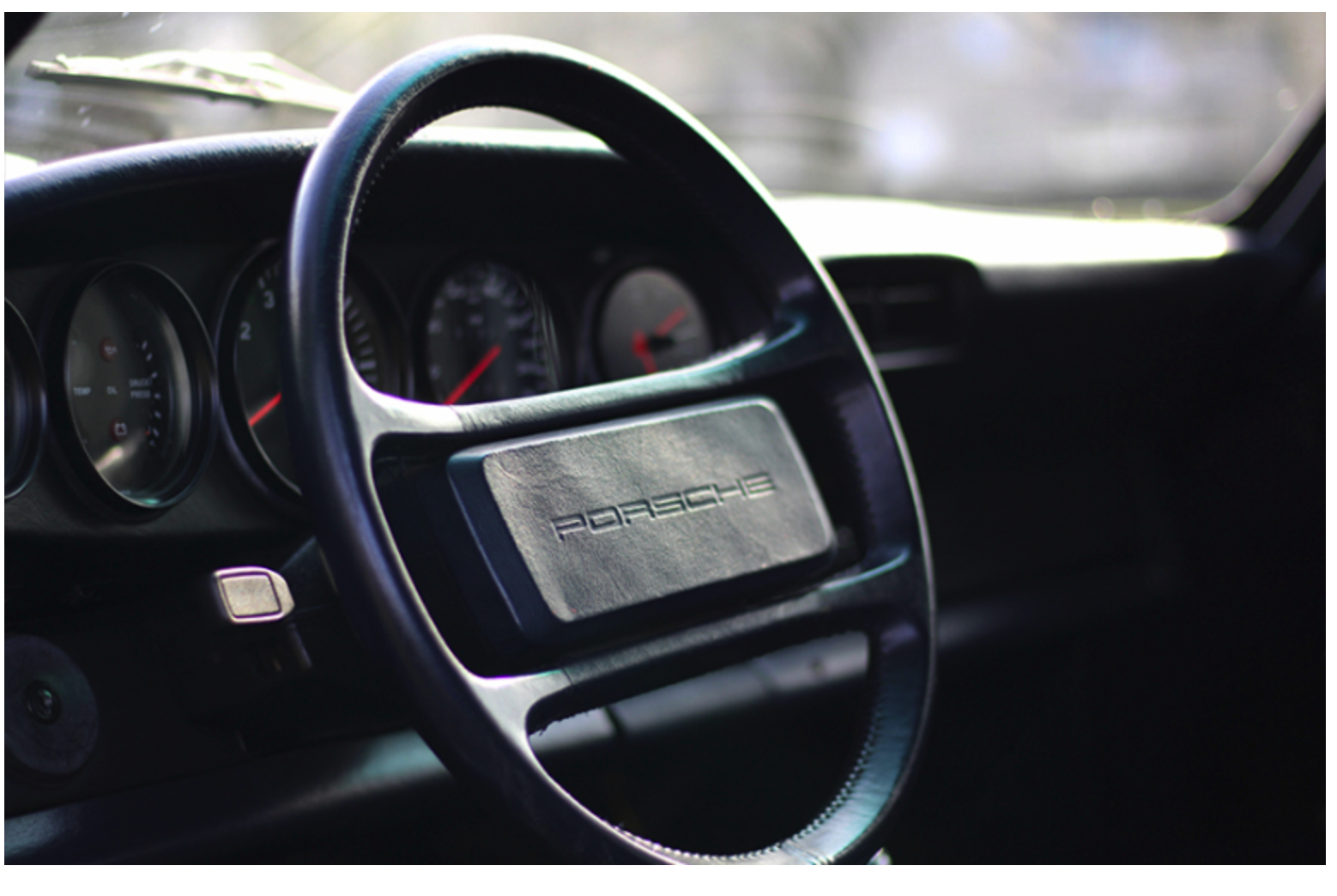 Nahaufnahme eines Oldtimer-Lenkrads mit dem Schriftzug Porsche in der Mitte, mit einem Teil des Armaturenbretts und Instrumentenanzeigen im Hintergrund. Die Beleuchtung hebt die strukturierte Oberfläche des Lenkrads hervor.