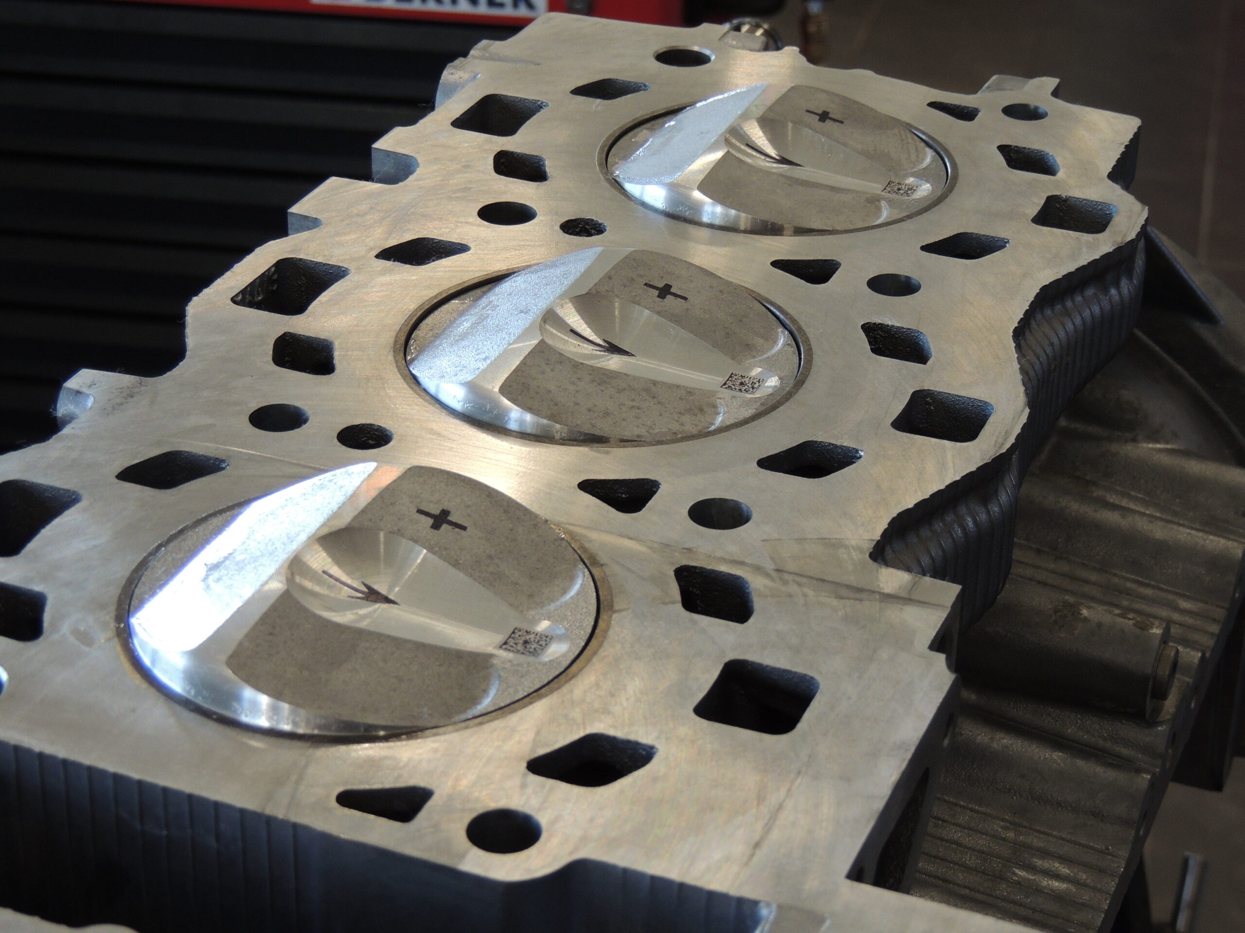 Nahaufnahme eines metallischen Motorblocks mit drei glänzenden Kolben, die jeweils mit einem Pluszeichen und einem Pfeil gekennzeichnet sind und sich in den jeweiligen Zylindern befinden.