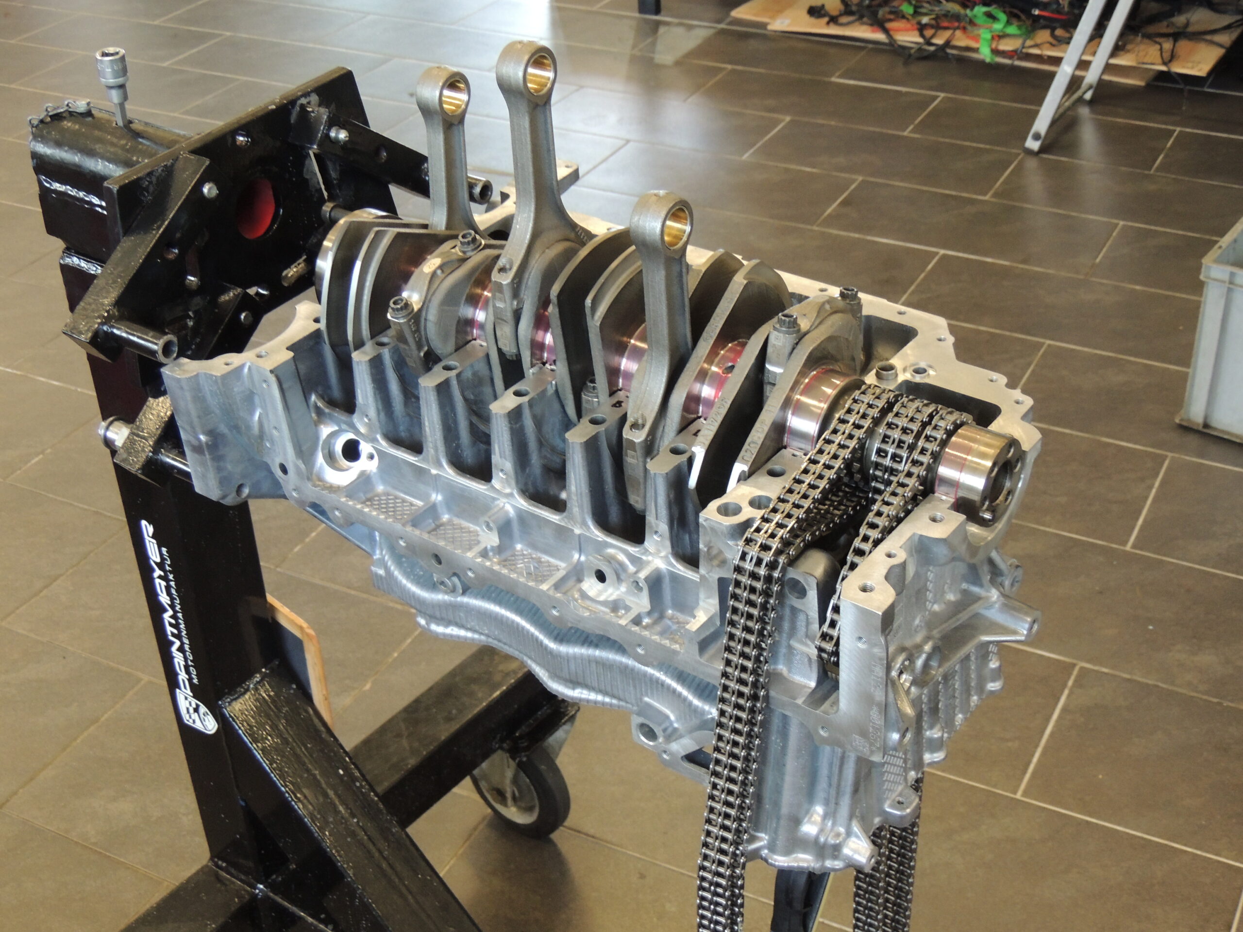 Ein teilweise zusammengebauter Motorblock auf einem Ständer mit Kurbelwelle, Pleuelstangen und Steuerketten, mit einem gekachelten Boden und verschiedenen Kabeln im Hintergrund.