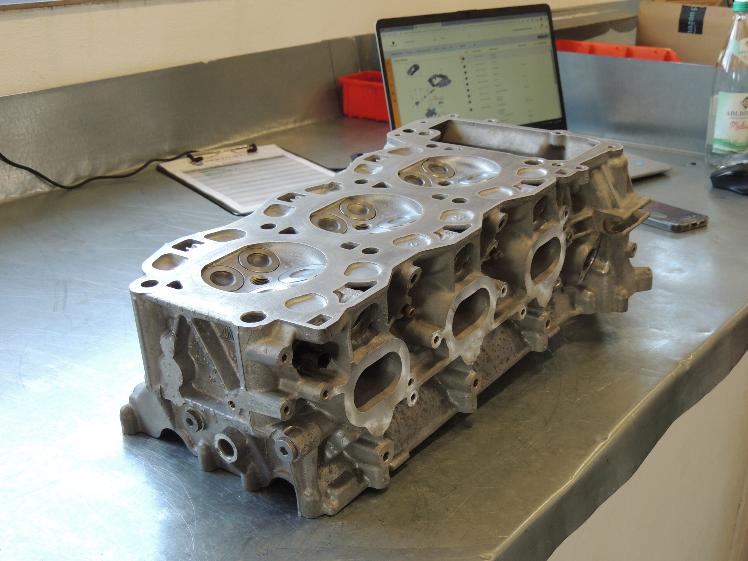 Ein Motorzylinderkopf aus Metall liegt auf einer Werkbank aus rostfreiem Stahl, im Hintergrund sind ein Laptop, ein Klemmbrett und eine Wasserflasche zu sehen.