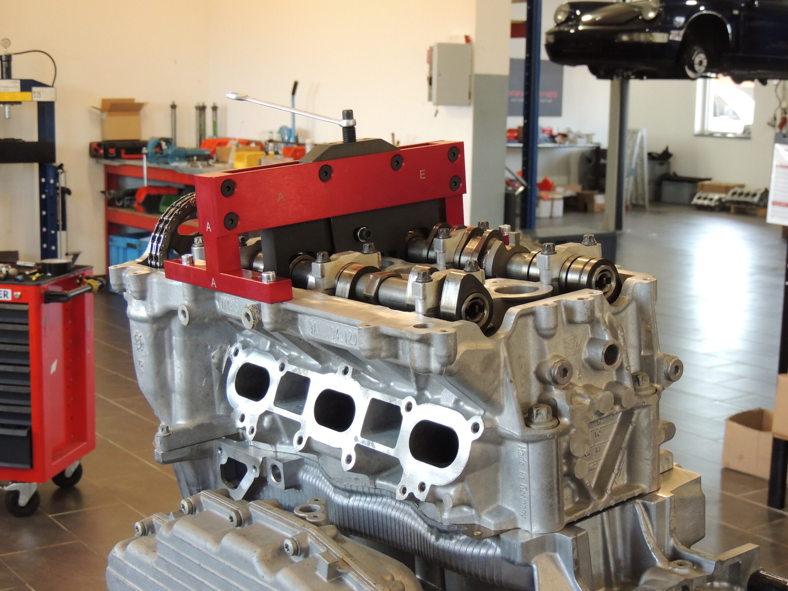 Nahaufnahme eines Motorblocks auf einem Ständer in einer Autowerkstatt, mit Werkzeugen und Geräten im Hintergrund und einem Auto, das auf einer Hebebühne angehoben wird.