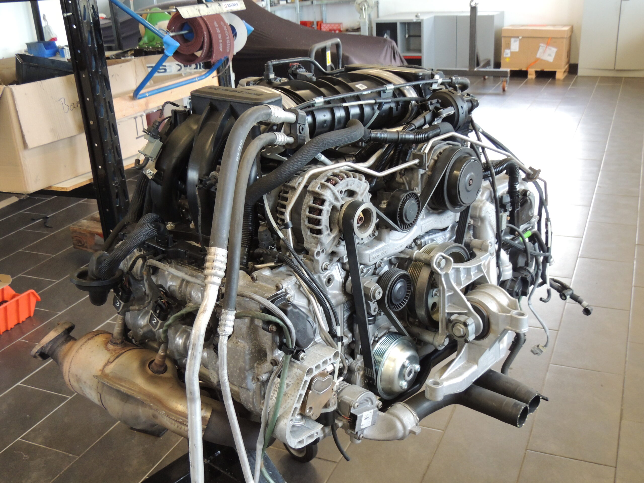 Ein moderner Automotor, der aus einem Fahrzeug ausgebaut wurde, liegt auf dem Boden einer Werkstatt, umgeben von Werkzeugen und Geräten. Verschiedene Schläuche, Riemen und Metallteile sind sichtbar und zeigen die komplexe Struktur des Motors.