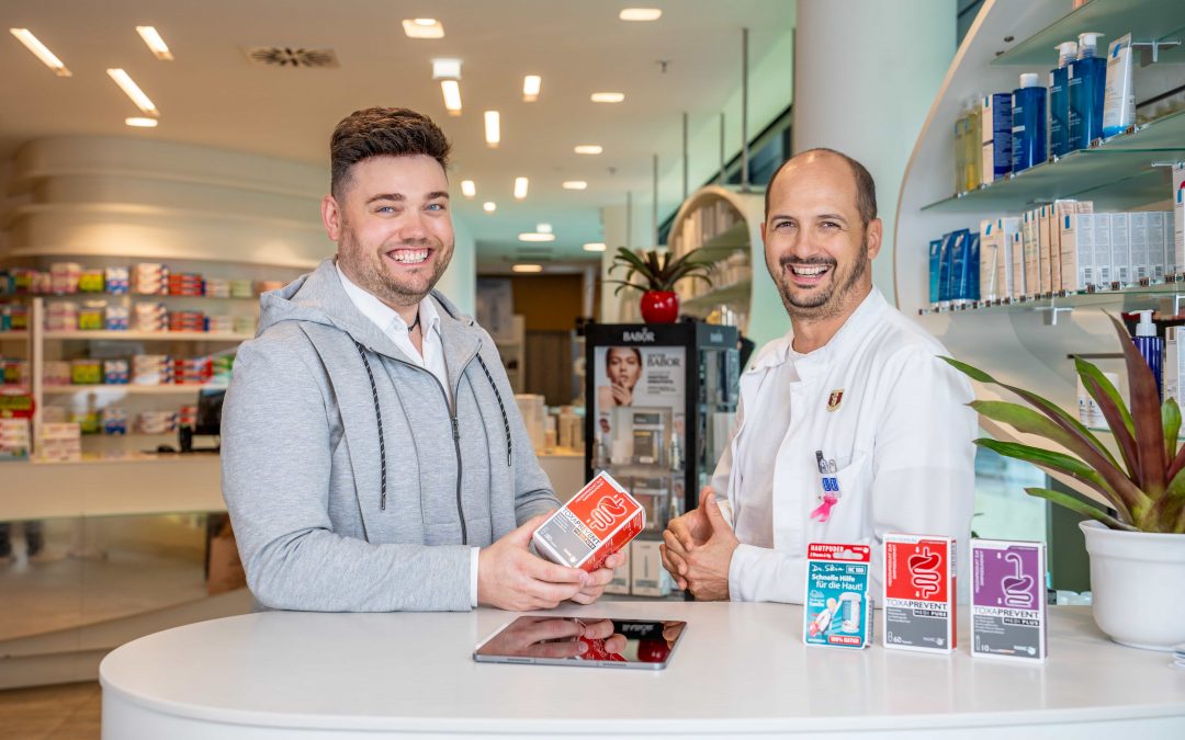 DominikKleeberg_sept24©invisions.at_1 Ein lächelnder Apotheker in einem weißen Kittel hilft einem Kunden mit einer Medikamentenschachtel an einem Apothekentresen. Im Hintergrund sind Regale mit Produkten und Informationsbroschüren zu sehen.