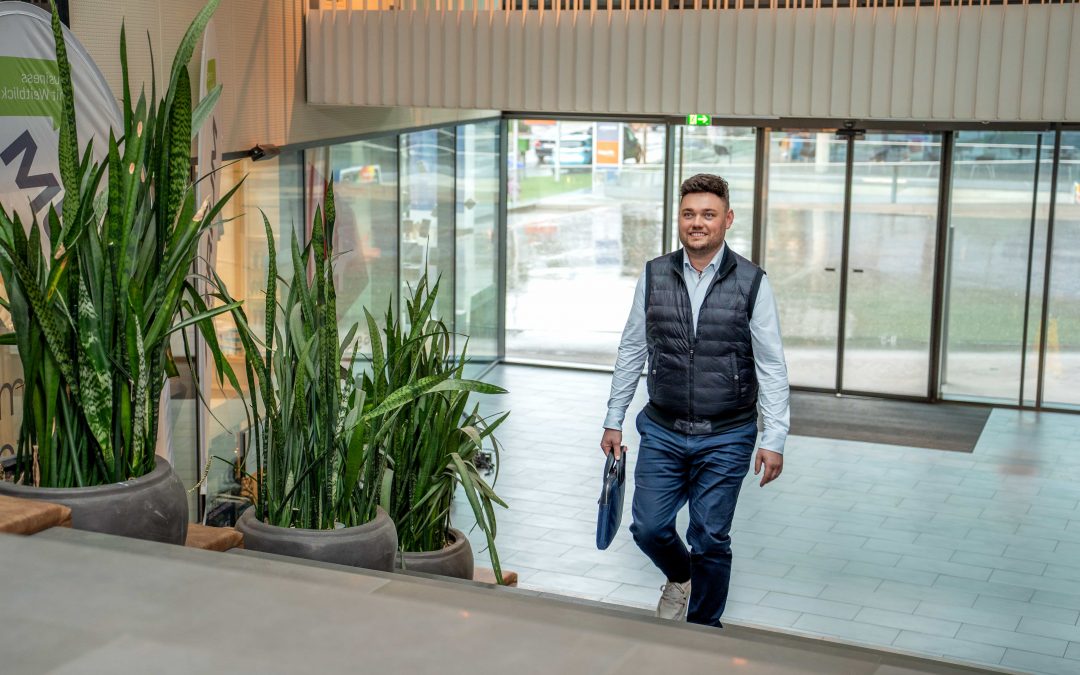 DominikKleeberg_sept24©invisions.at_105 Ein Mann in blauer Weste und Jeans geht eine Innentreppe in der Nähe großer Topfpflanzen hinauf und hält einen blauen Ordner in der Hand. Im Hintergrund sind Glastüren und ein Außenbereich zu sehen, was auf ein Büro- oder Geschäftsgebäude hindeutet.