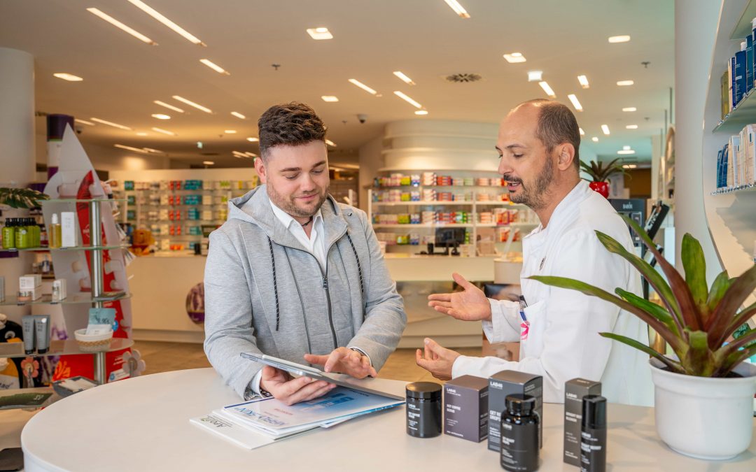 DominikKleeberg_sept24©invisions.at_42 Zwei Männer stehen an einem Apothekentresen; der eine im weißen Kittel gestikuliert, während der andere, in legerer Kleidung, auf ein Tablet schaut. Auf dem Tresen liegen Hautpflegeprodukte und Papiere, und im Hintergrund sind Regale mit Produkten aufgebaut.