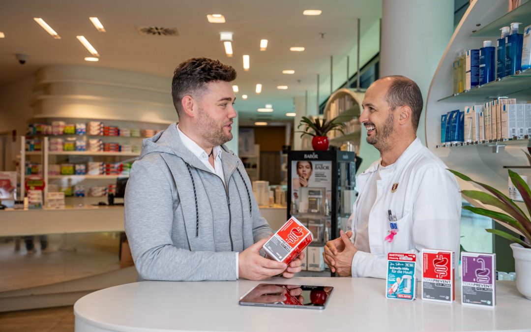 DominikKleeberg_sept24©invisions.at_65 Ein Mann in Freizeitkleidung berät sich mit einem Apotheker im weißen Kittel an einem Apothekentresen, der eine Medikamentenschachtel in der Hand hält und lächelt, während sie sich unterhalten. Im Hintergrund sind Regale mit Produkten zu sehen.