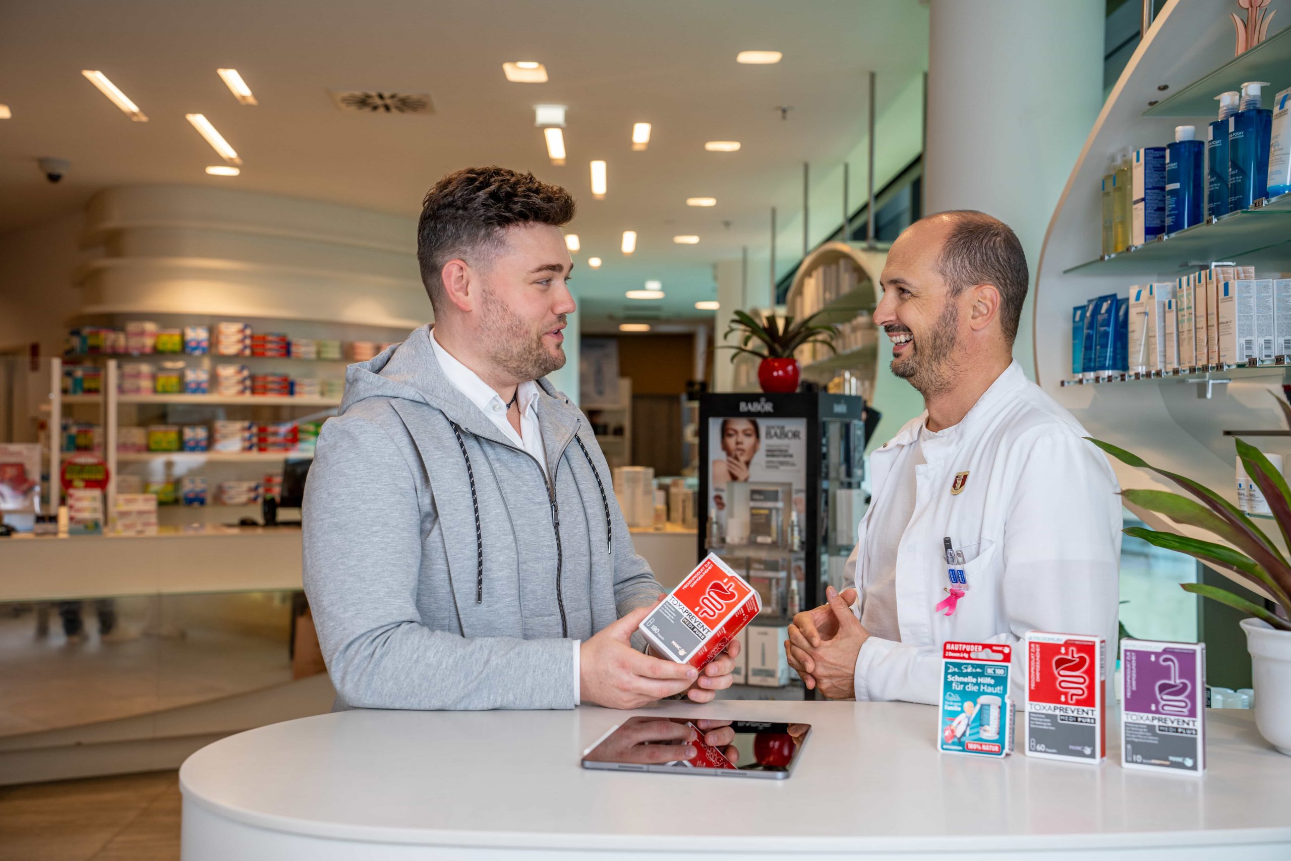 Ein Mann in Freizeitkleidung berät sich mit einem Apotheker im weißen Kittel an einem Apothekentresen, der eine Medikamentenschachtel in der Hand hält und lächelt, während sie sich unterhalten. Im Hintergrund sind Regale mit Produkten zu sehen.