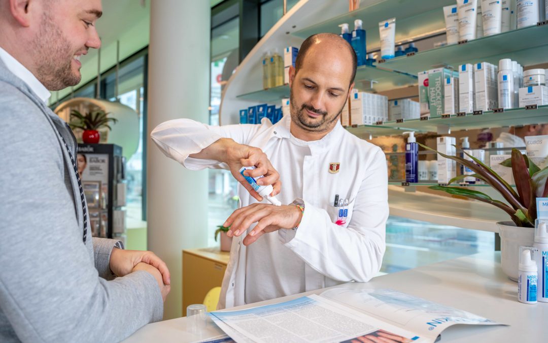 DominikKleeberg_sept24©invisions.at_73 Ein Apotheker in einem weißen Kittel demonstriert einer Kundin an der Apothekentheke ein Hautpflegeprodukt auf seiner Hand, während im Hintergrund Regale mit Produkten zu sehen sind.