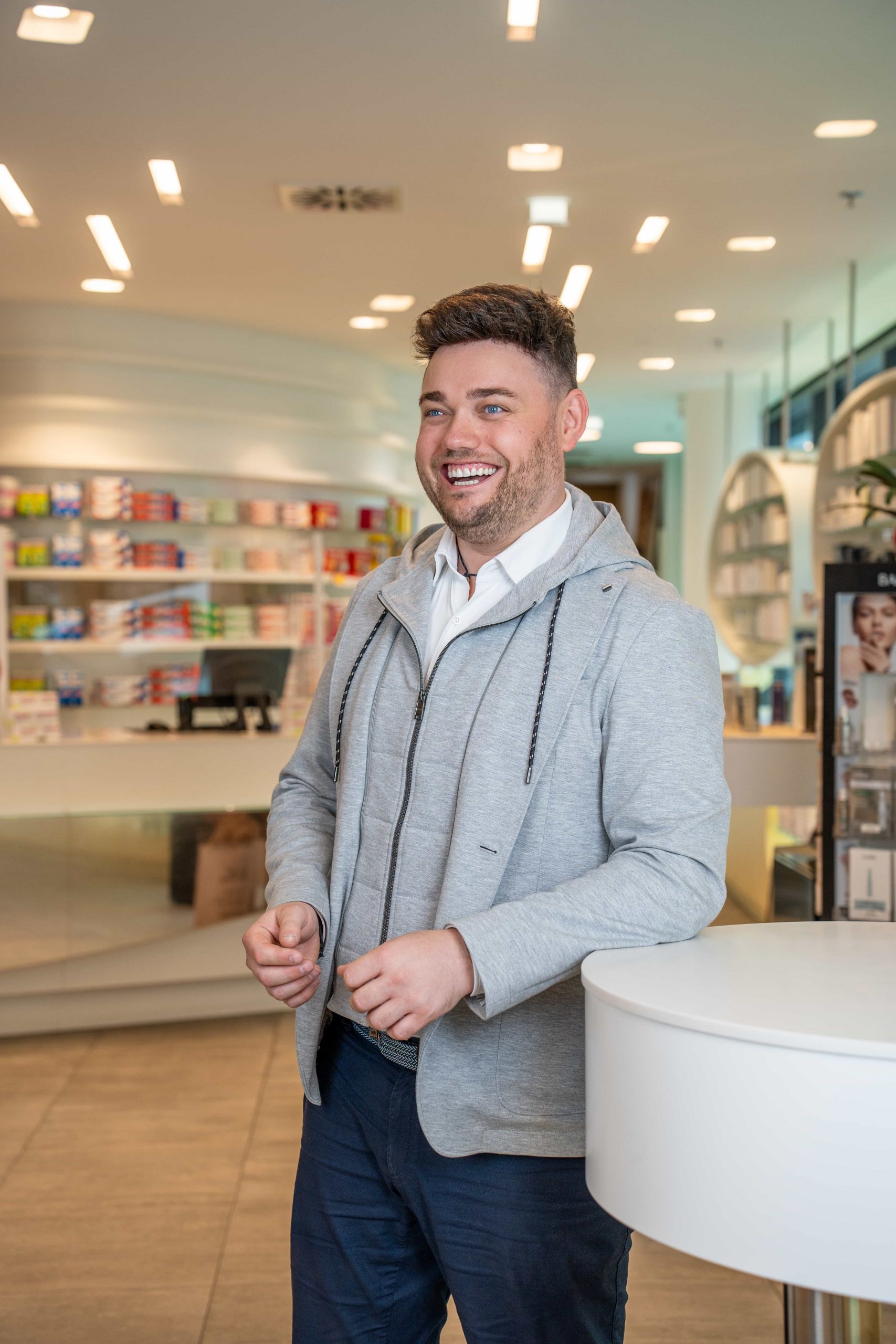 Ein lächelnder Mann in einem hellgrauen Kapuzenpulli und einer dunklen Hose steht in einer hell erleuchteten Apotheke oder einem Geschäft, mit Regalen voller Produkte und moderner Einrichtung im Hintergrund.