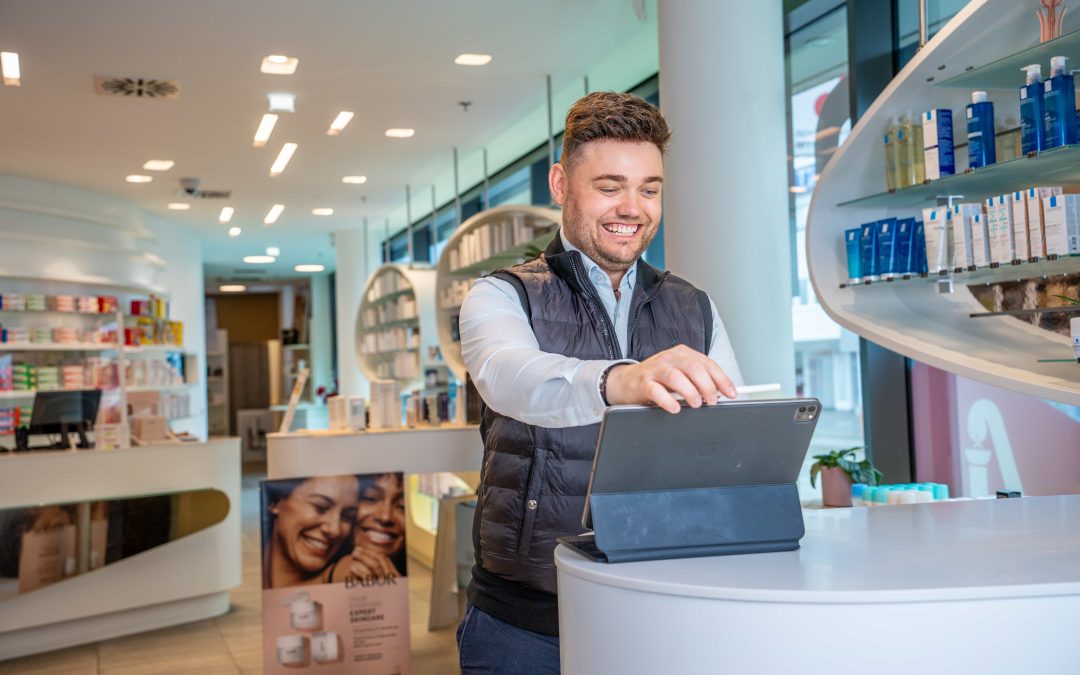 DominikKleeberg_sept24©invisions.at_90 Ein lächelnder Mann in einer Weste benutzt ein Tablet auf einem Tresen in einer hellen, modernen Apotheke mit Regalen voller Produkte und Hautpflegewerbung im Hintergrund.