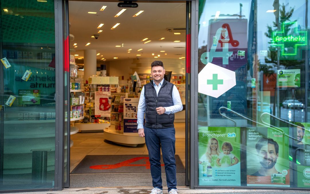DominikKleeberg_sept24©invisions.at_98 Ein Mann mit schwarzer Weste, weißem Hemd und blauen Jeans steht lächelnd vor einem modernen Apothekeneingang mit Glastüren und einem grünen Kreuz. Im Inneren sind Apothekenregale und Produkte zu sehen.