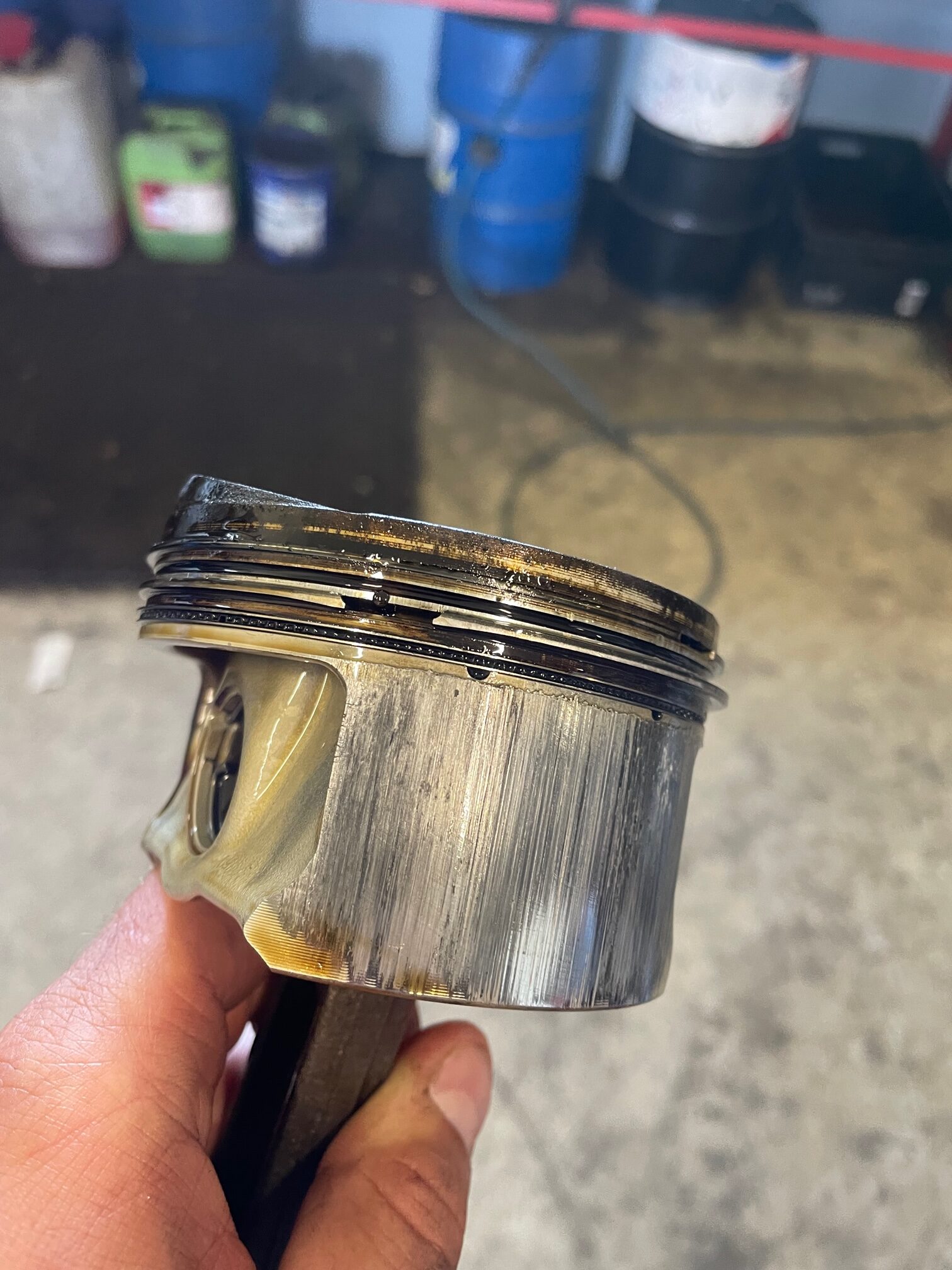 Eine Hand hält einen abgenutzten Motorkolben mit sichtbaren Riefen, Verfärbungen und Kohlenstoffablagerungen in einer Garage mit Ölfässern und Geräten im Hintergrund.