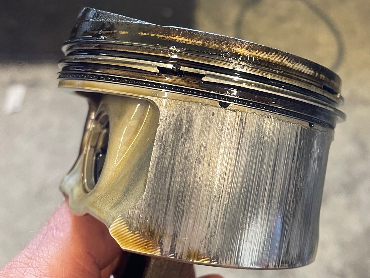 Nahaufnahme eines beschädigten Motorkolbens mit sichtbaren vertikalen Riefen und Verfärbungen an der Seite, die auf Verschleiß und mögliche Fehlfunktionen hinweisen.