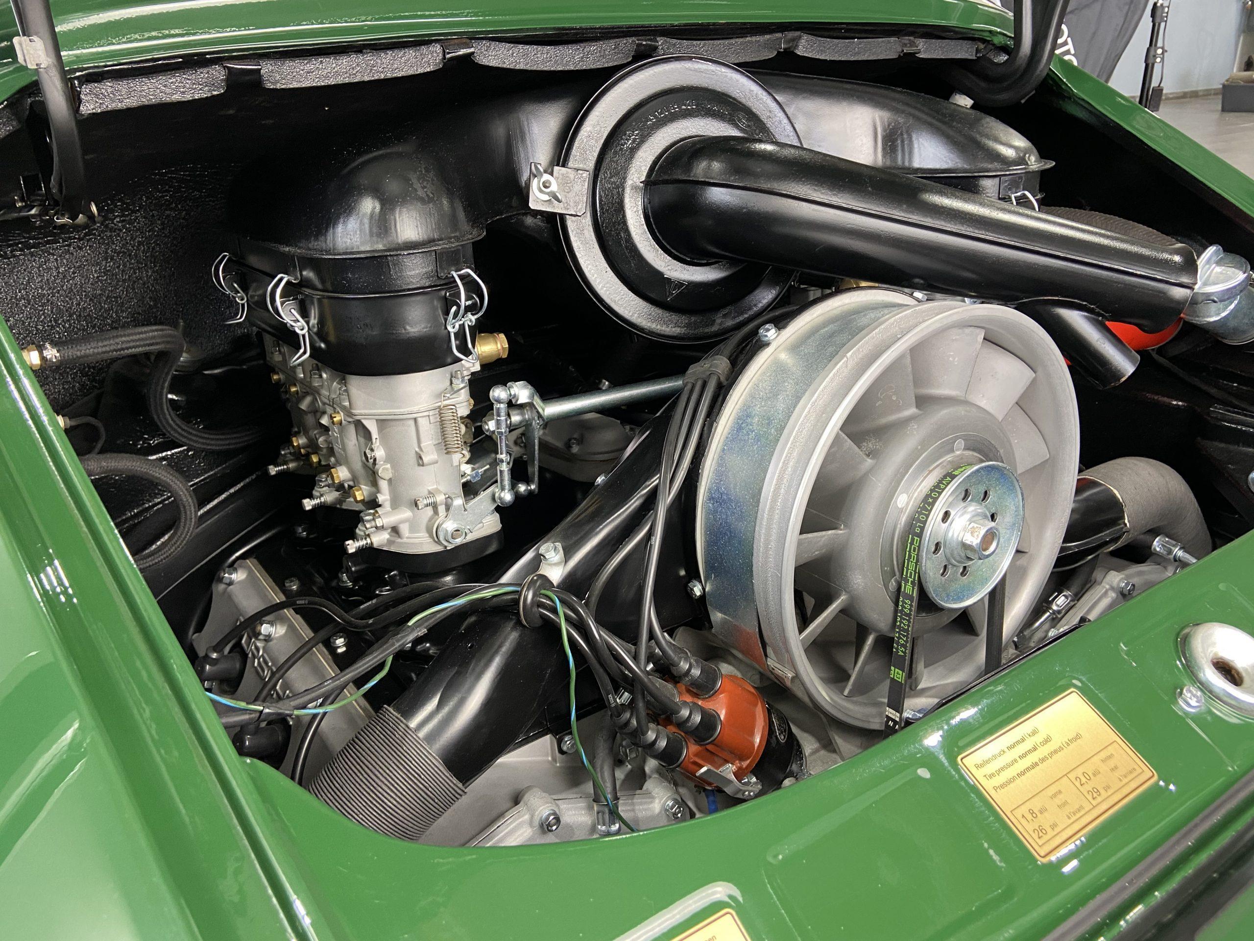 Nahaufnahme eines Oldtimer-Motors in einem grünen Motorraum, mit detaillierten Komponenten wie Luftfilter, Lüfter, Kabeln und goldenen Hinweisschildern am Fahrzeug.