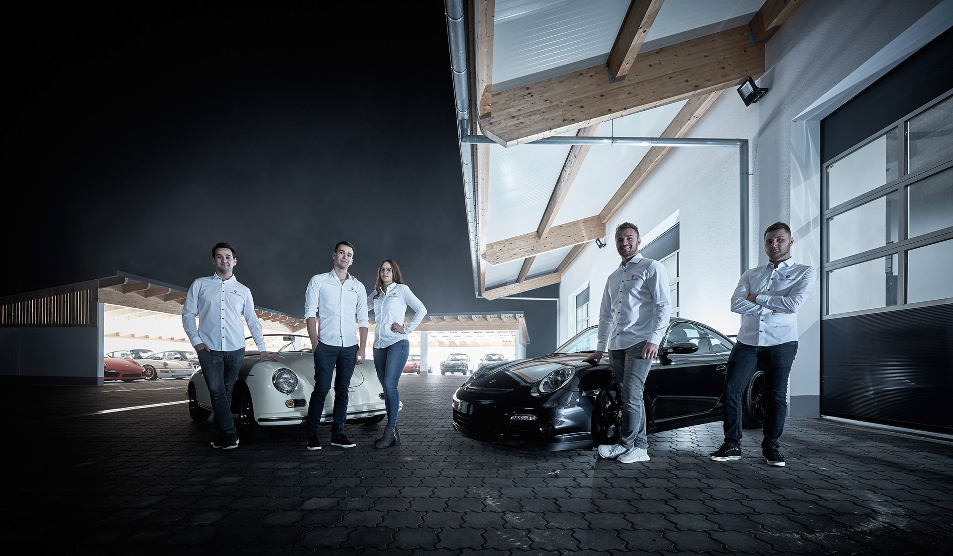 Fünf Personen in weißen Hemden und dunklen Hosen stehen selbstbewusst vor zwei luxuriösen Sportwagen, einem weißen und einem schwarzen, unter einem modernen, holzüberdachten Carport mit großen Fenstern.