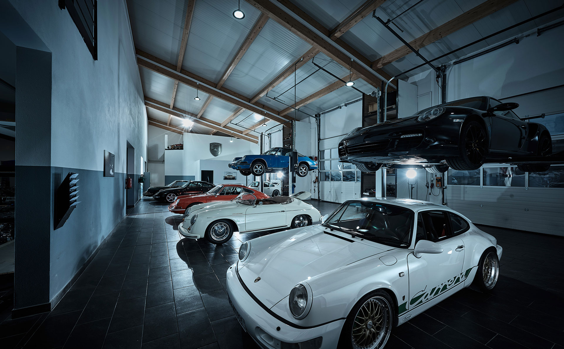 Eine schicke, moderne Garage mit mehreren Klassikern und Sportwagen, darunter weiße, rote und schwarze Porsche-Modelle. Ein schwarzer Wagen ist mit einem Aufzug hochgefahren, und der Raum hat hohe Decken mit sichtbaren Holzbalken.