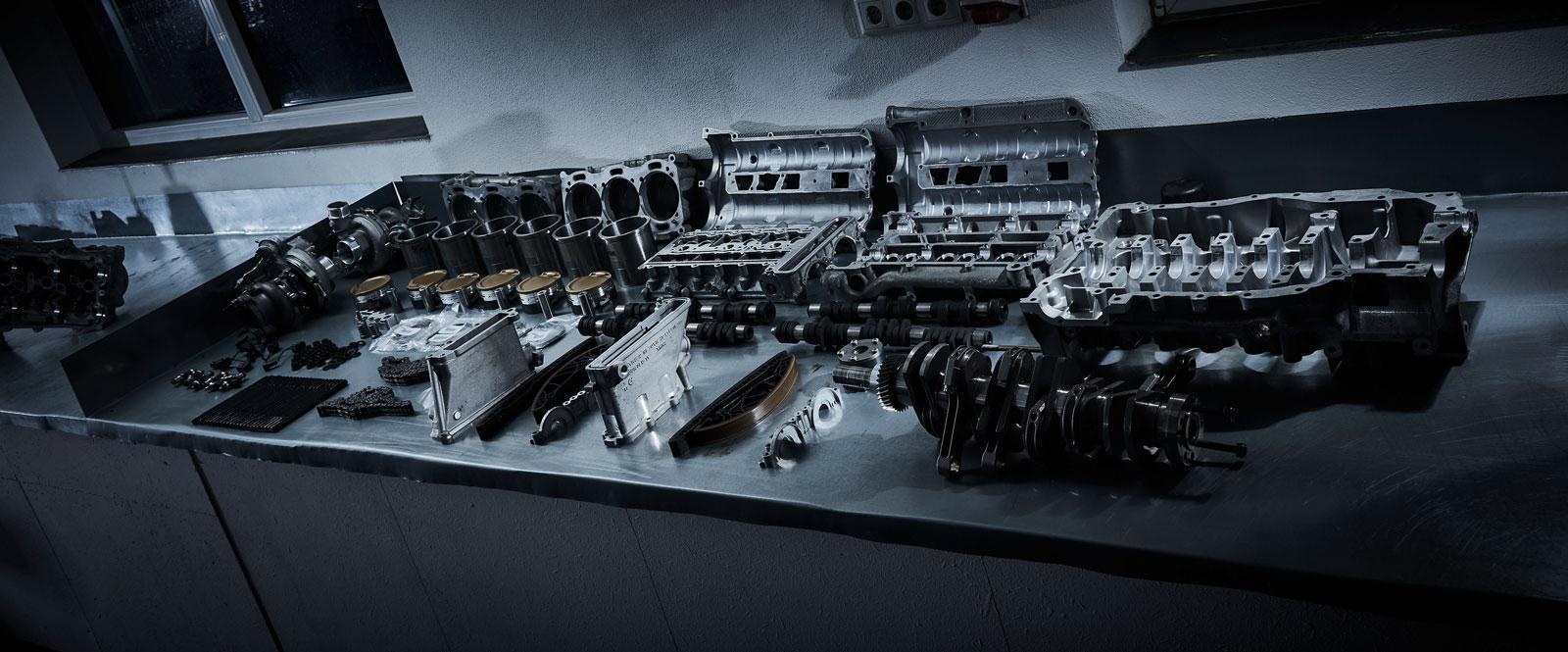 Auf einer metallenen Werkbank in einer schwach beleuchteten Werkstatt sind verschiedene zerlegte Motorenteile, darunter Kolben, Nockenwellen, Kurbelwellen und Zylinderköpfe, fein säuberlich angeordnet.