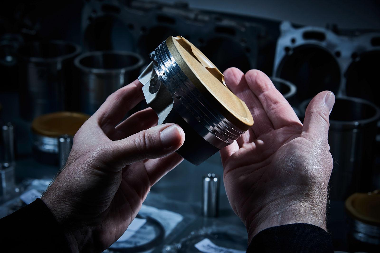 Eine Person hält einen Metallkolben mit einer goldenen Spitze und untersucht dessen Details. Andere Motorteile und Kolben sind unscharf im Hintergrund auf einer Werkbank zu sehen. Die Beleuchtung hebt das Objekt und die Hände hervor.