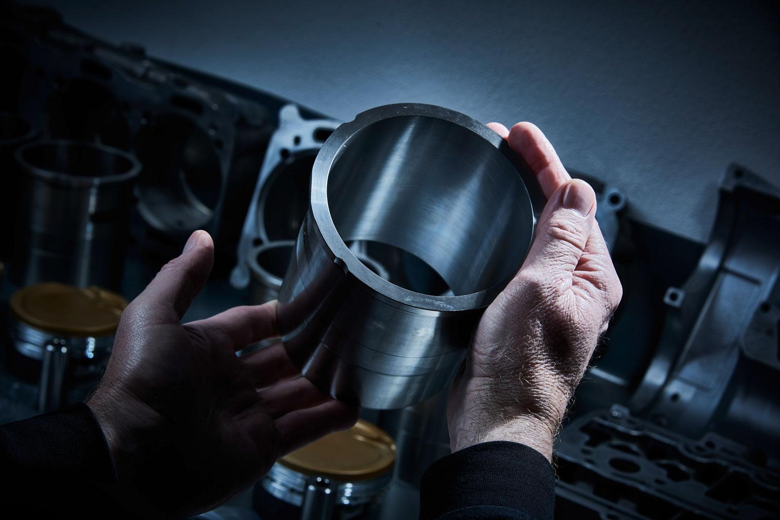 Die Hände einer Person halten einen großen, glänzenden Metallzylinder, möglicherweise ein Motorteil, während andere Metallteile und Maschinen im Hintergrund bei dramatischer Beleuchtung verschwimmen.
