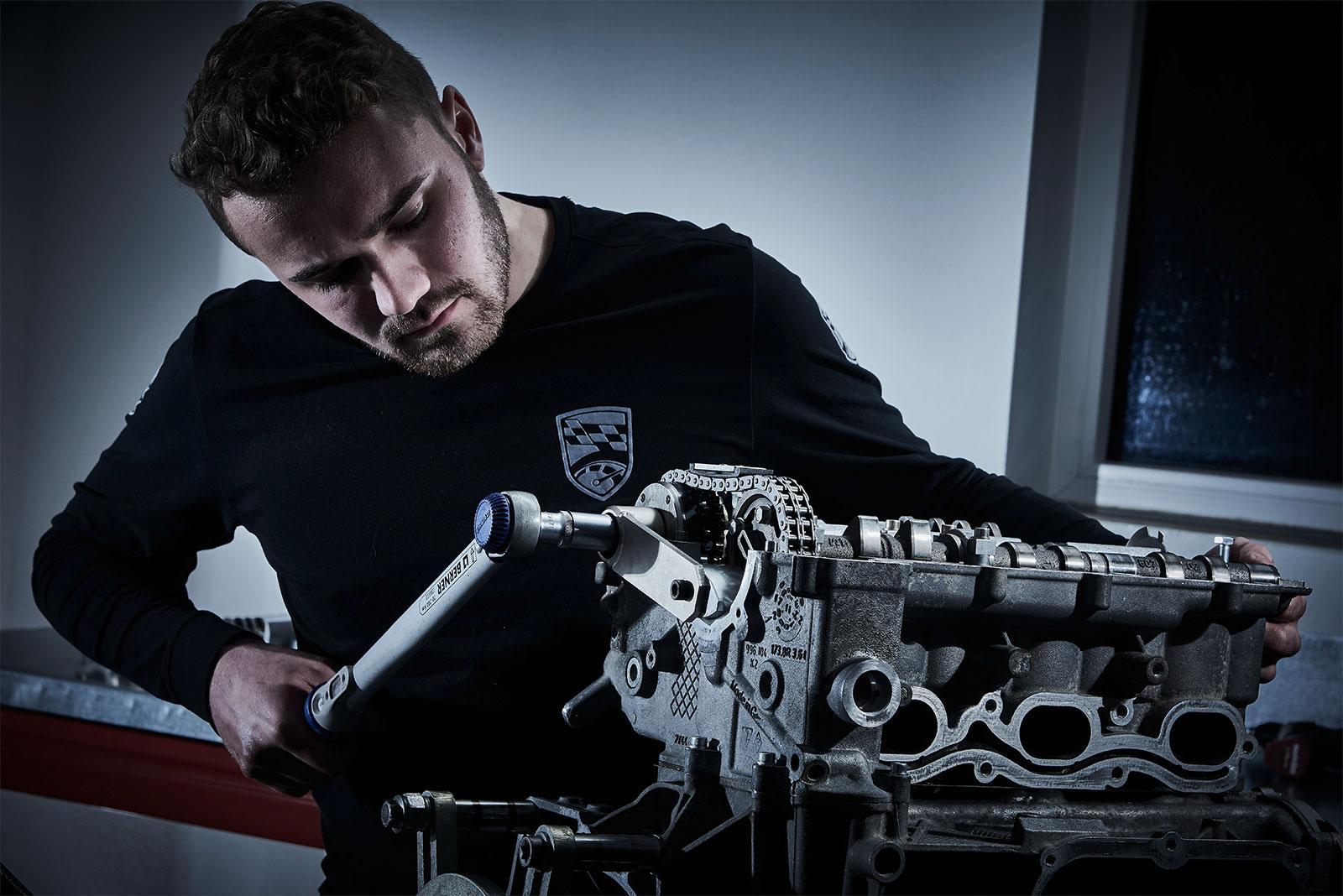 Ein Mann in einem dunklen Hemd arbeitet mit einem Drehmomentschlüssel an einem freiliegenden Motorteil in einer schwach beleuchteten Werkstatt.