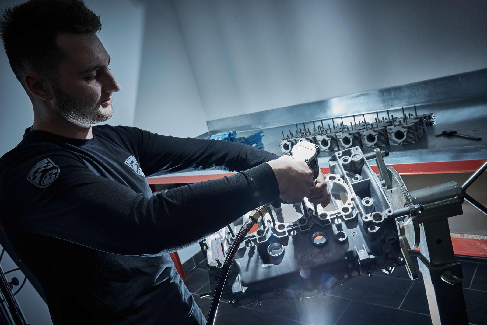 Ein Mann in einem schwarzen Hemd arbeitet mit einem Elektrowerkzeug an einem Motorblock aus Metall in einer Werkstatt, während im Hintergrund Motorteile und Werkzeuge auf einer Metallwerkbank zu sehen sind.
