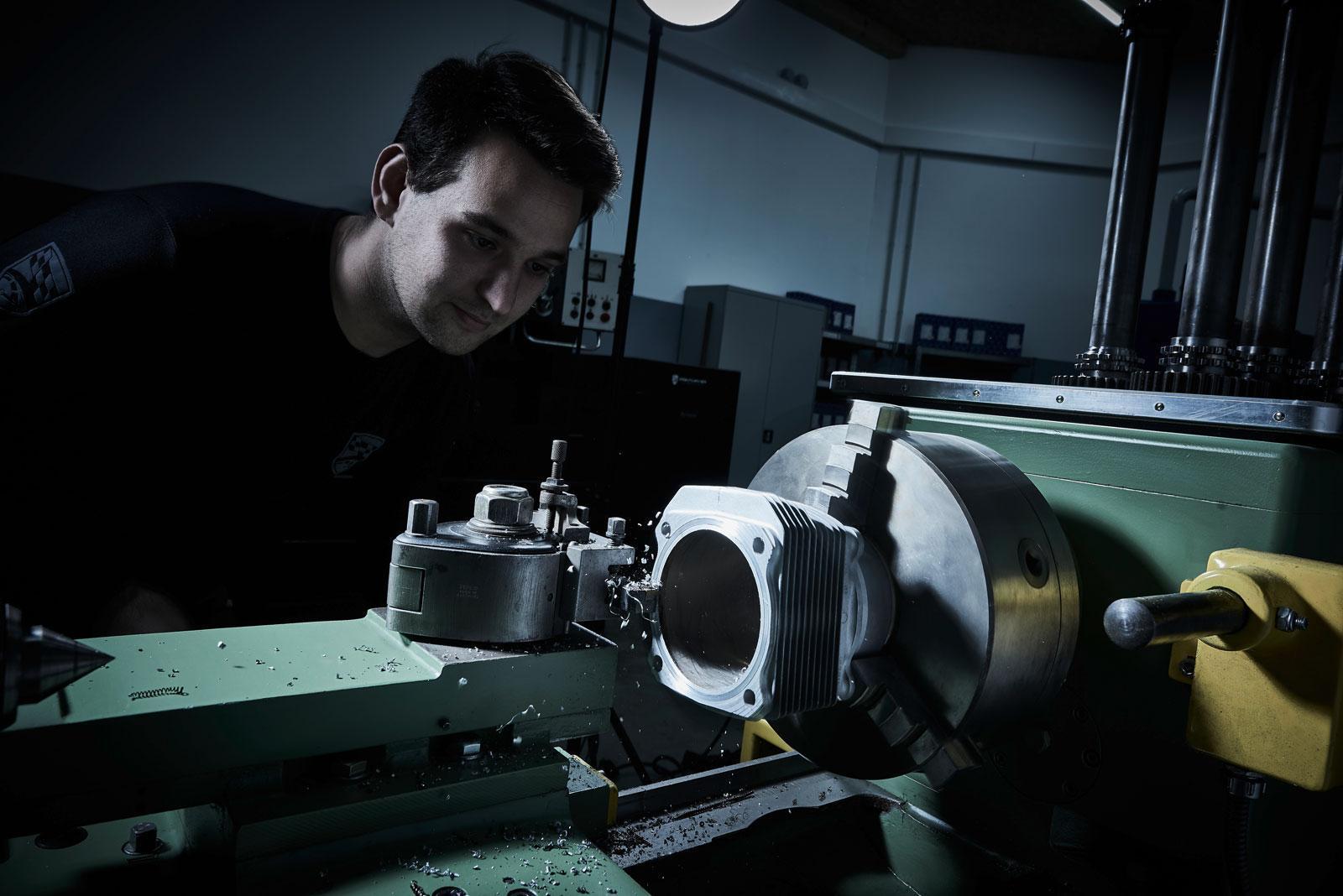 Ein Mann bedient eine grüne industrielle Drehmaschine in einer schwach beleuchteten Werkstatt und beobachtet genau, wie ein Metallwerkstück bearbeitet wird. Auf der Oberfläche der Maschine sind Metallspäne verstreut.