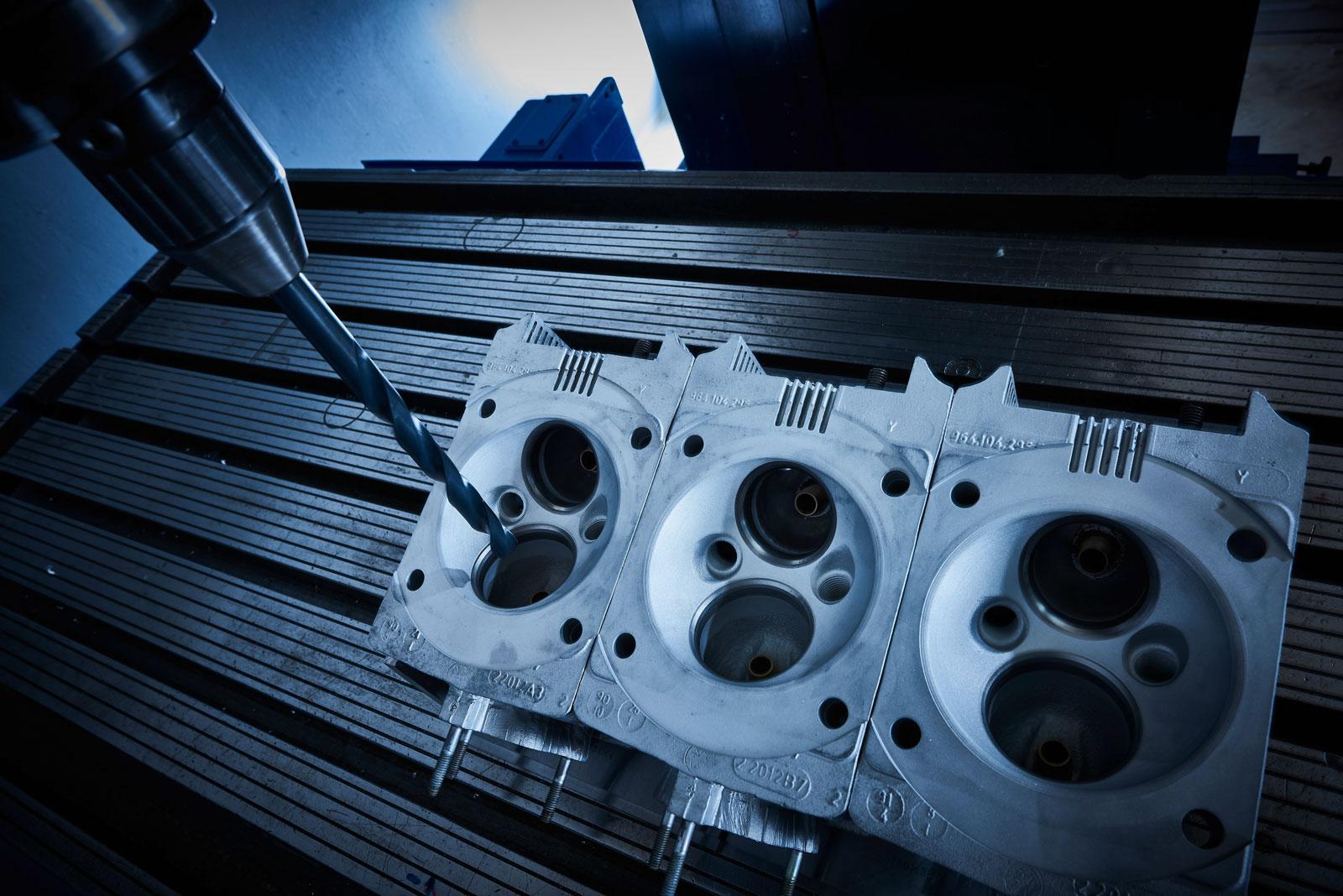 Nahaufnahme einer CNC-Maschine, die einen Zylinderkopf aus Metall bohrt, wobei drei Verbrennungskammern sichtbar sind, auf einem dunklen industriellen Arbeitstisch mit blauer Beleuchtung.