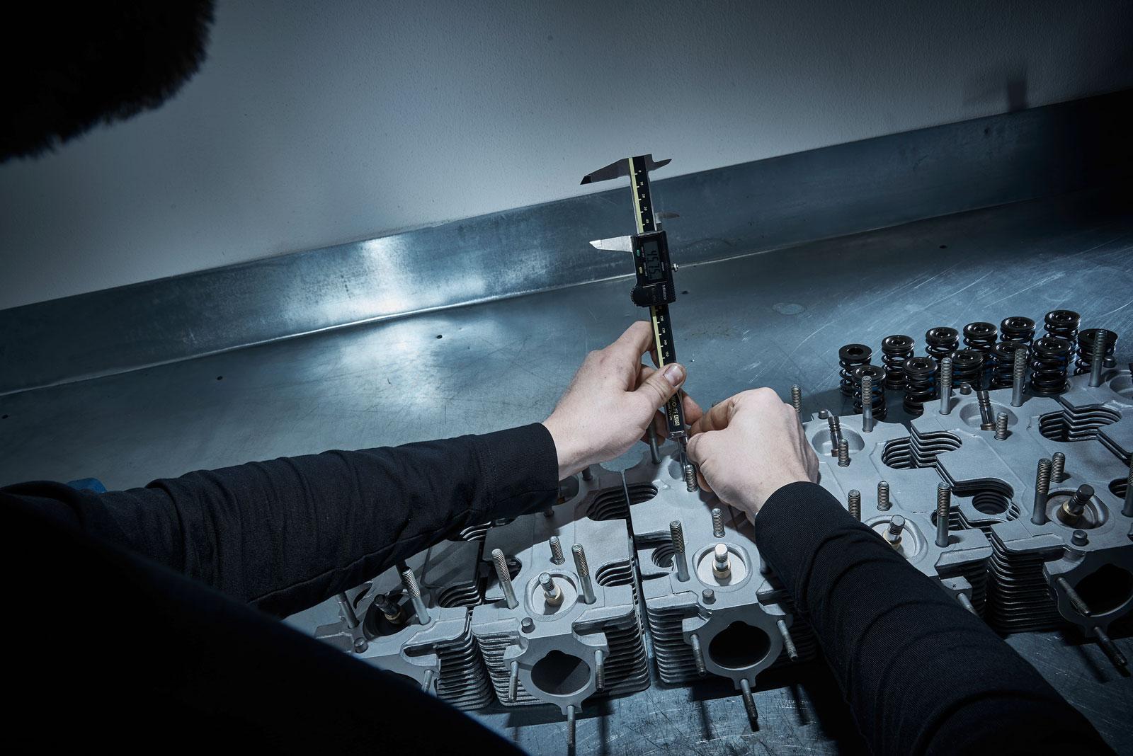Eine Person misst mit einem digitalen Messschieber ein Metallteil an einer Zylinderkopfbaugruppe, während andere Motorteile und Federn auf einer Metallwerkbank unter fokussierter Beleuchtung sichtbar sind.