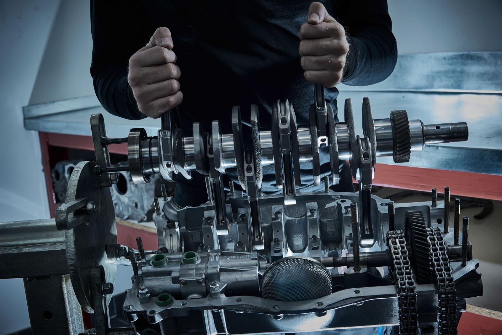 Eine Person, die eine polierte Metallkurbelwelle über einem teilweise zerlegten Motorblock in einer Werkstatt hält, wobei um die Maschine herum Zahnräder und Ketten zu sehen sind.