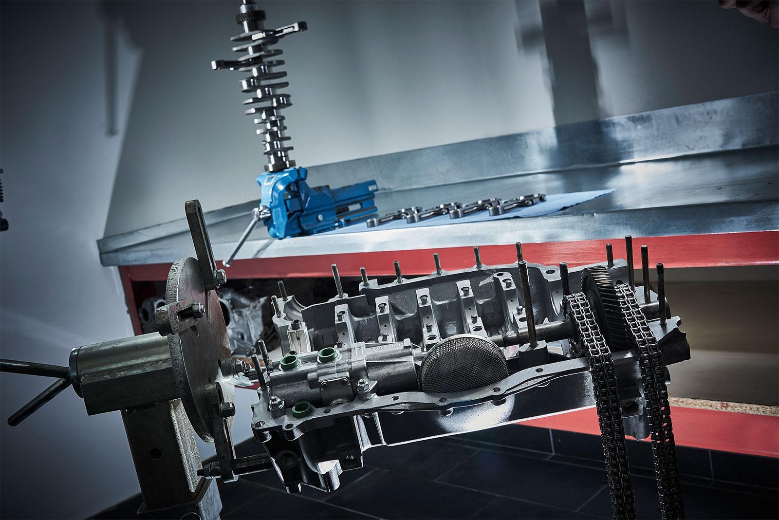 Nahaufnahme eines zerlegten Motorblocks, der auf einem Ständer in einer Werkstatt montiert ist, mit verschiedenen Motorteilen, Werkzeugen und einem blauen Behälter auf einer Werkbank im Hintergrund.